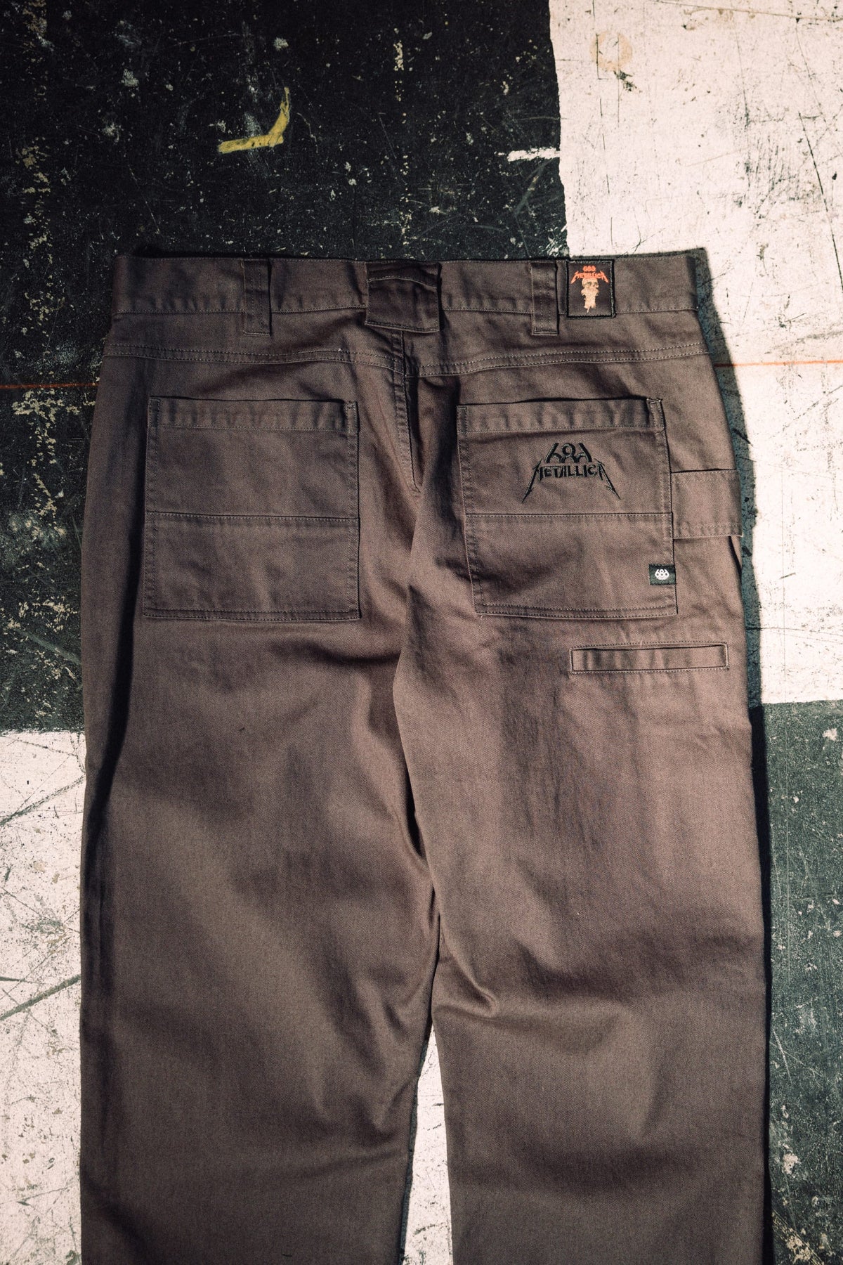 Dark brown 686 Men’s Unwork® Tradesman Pant - Relaxed Fit featuring a Metallica logo embroidered on the right back pocket and a small red logo patch on the waistband.