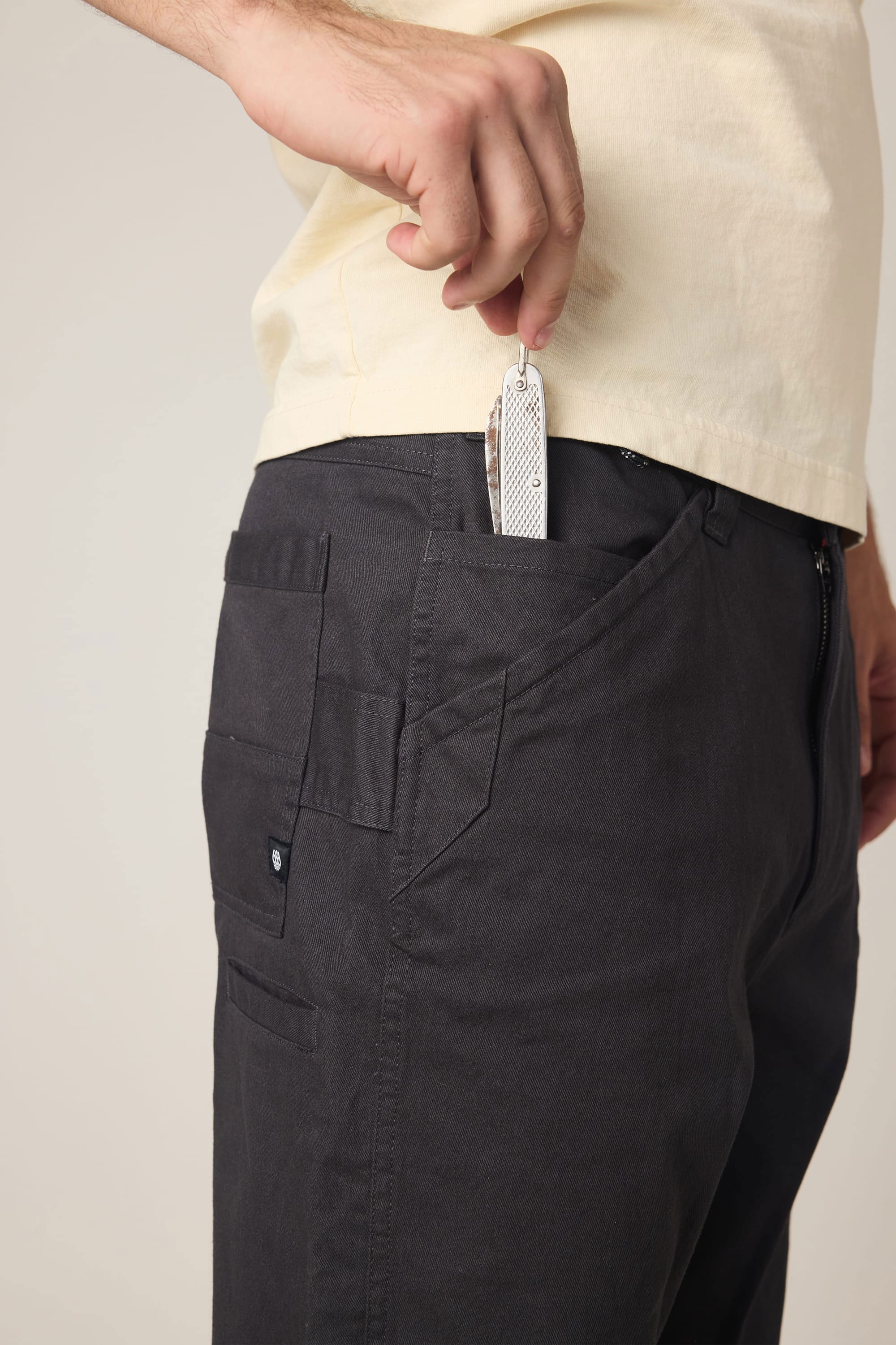 A person wearing a 686 Men's Unwork® Tradesman Pant - Relaxed Fit places a silver pocket knife into their front pants pocket.