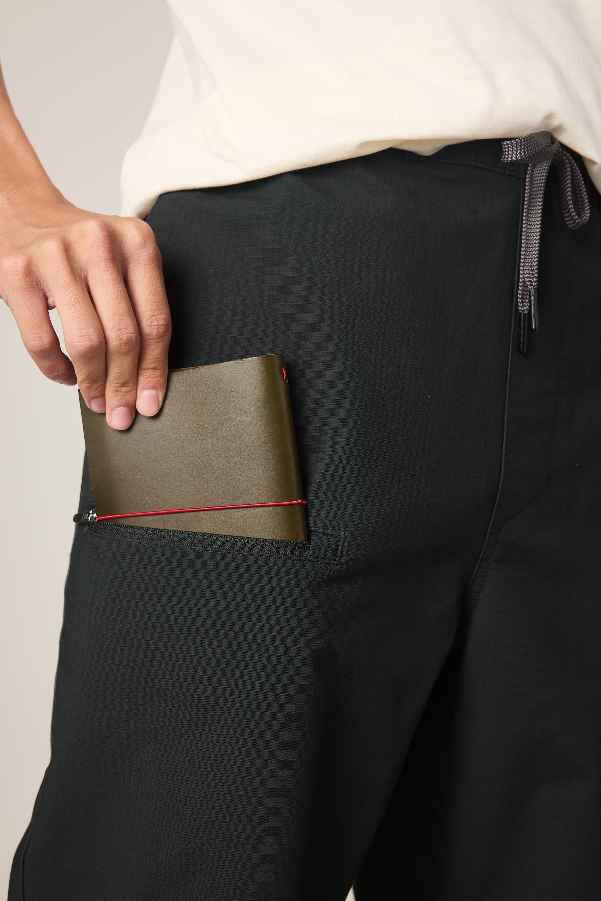 Dressed in a white shirt and 686 Men's Free Loader Pant - Loose Fit by 686, a person places a green wallet into the pant's zippered side pocket.