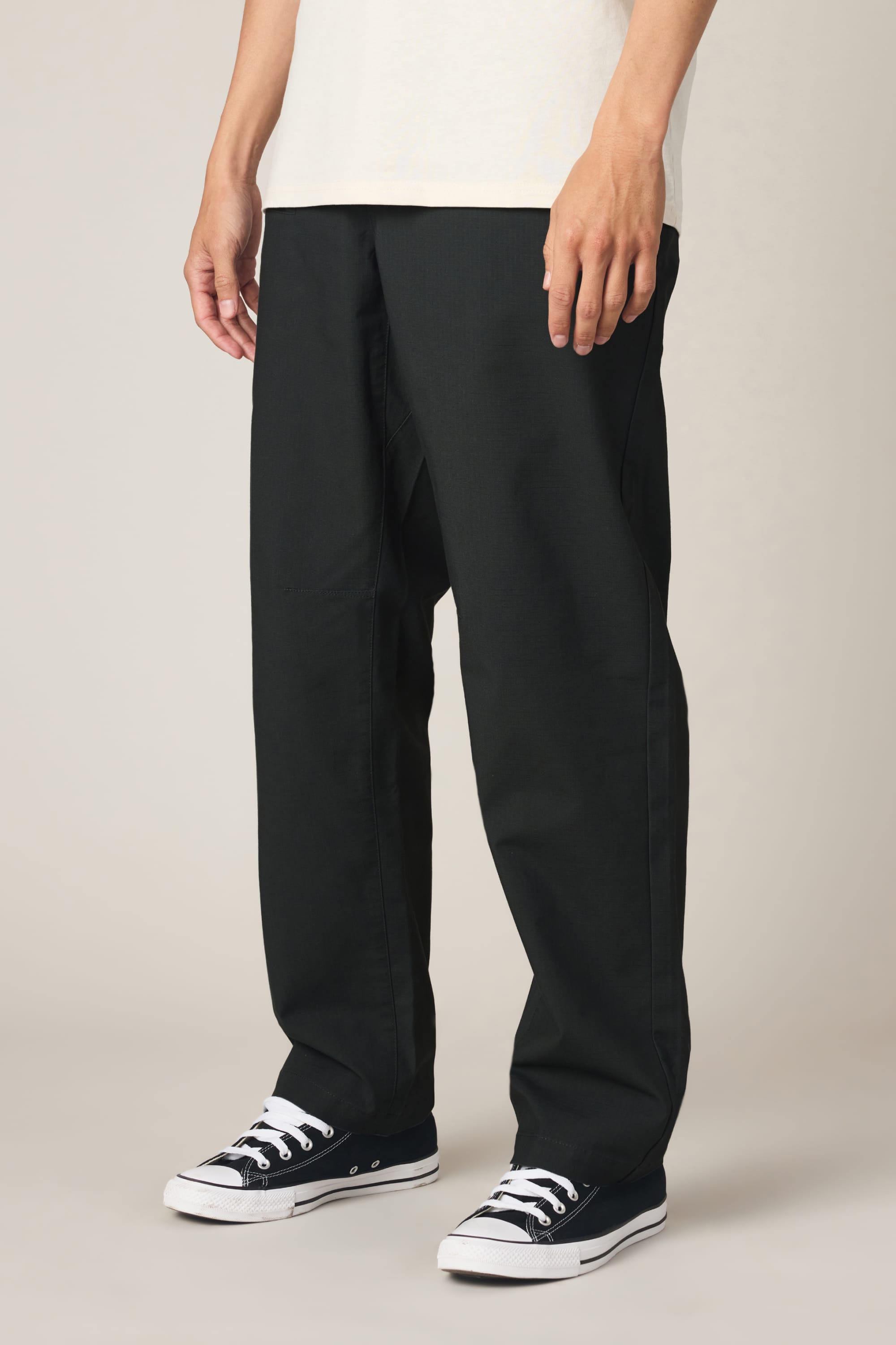 A person in 686 Men's Free Loader Pant - Loose Fit, a white shirt, and black low-top sneakers with white soles stands against a plain beige background.