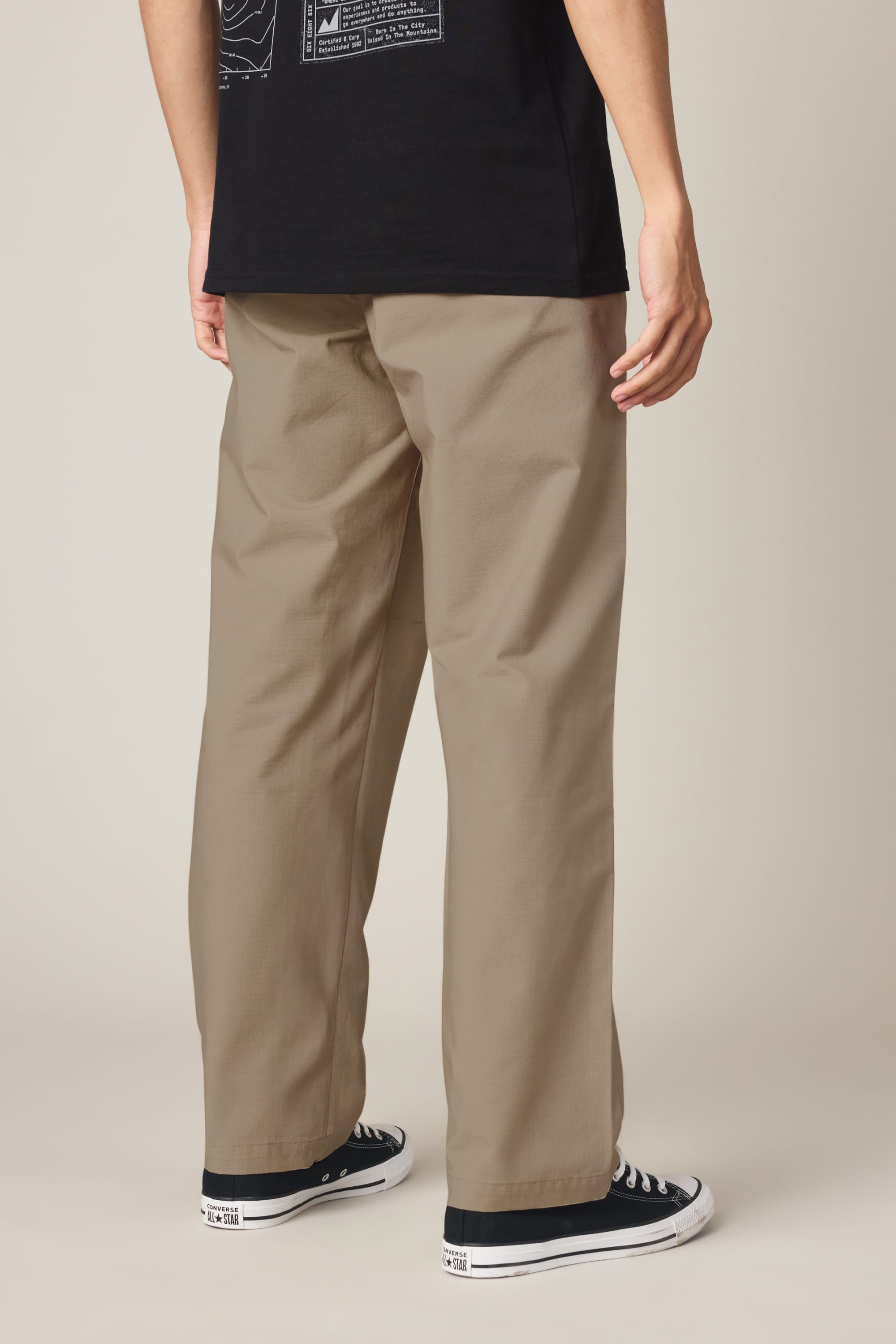 A person stands with their back to the camera, wearing a black t-shirt, black low-top sneakers with white soles, and the 686 Men's Free Loader Pant - Loose Fit by 686. The background is plain and light-colored.