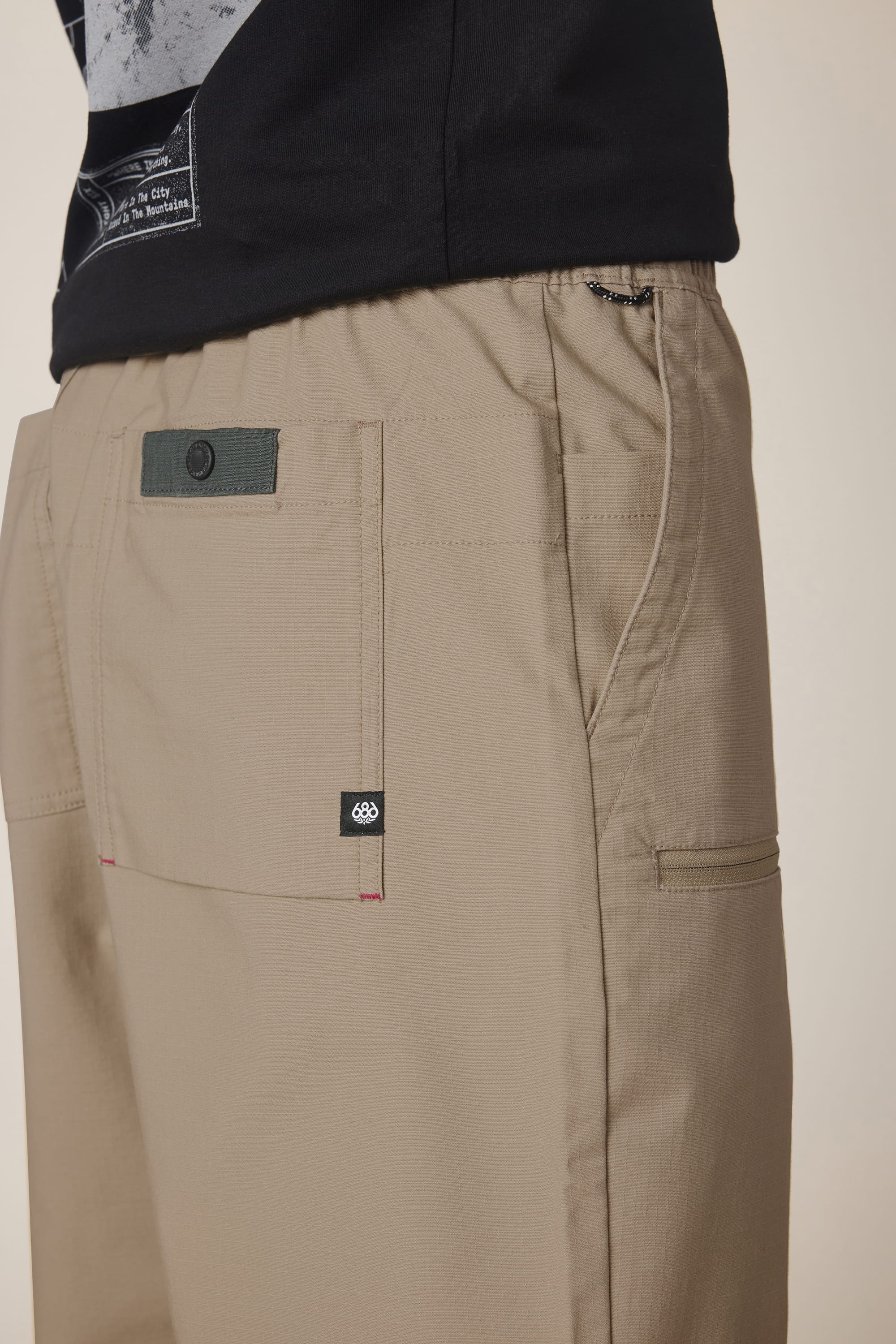 A close-up side view of the 686 Men's Free Loader Pant - Loose Fit, featuring large front pockets, a small black tag, and an elastic waistband, paired with a black T-shirt.