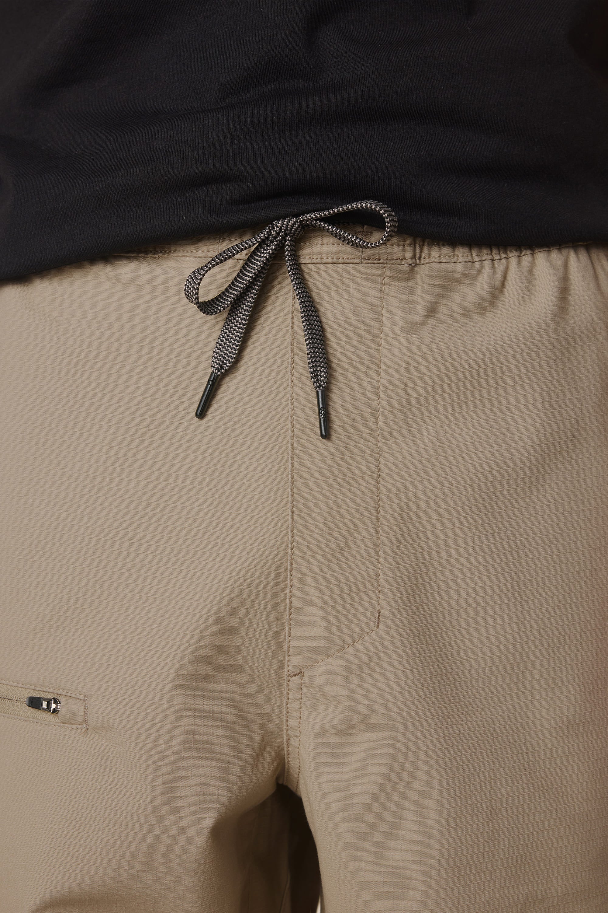 Close-up of a person wearing the 686 Men's Free Loader Pant - Loose Fit in light brown nylon, featuring a centered knot on the gray and black drawstring above the waistband. A zippered pocket is partially visible on the left side, highlighting the functional design by 686. The pants are paired with a black shirt.