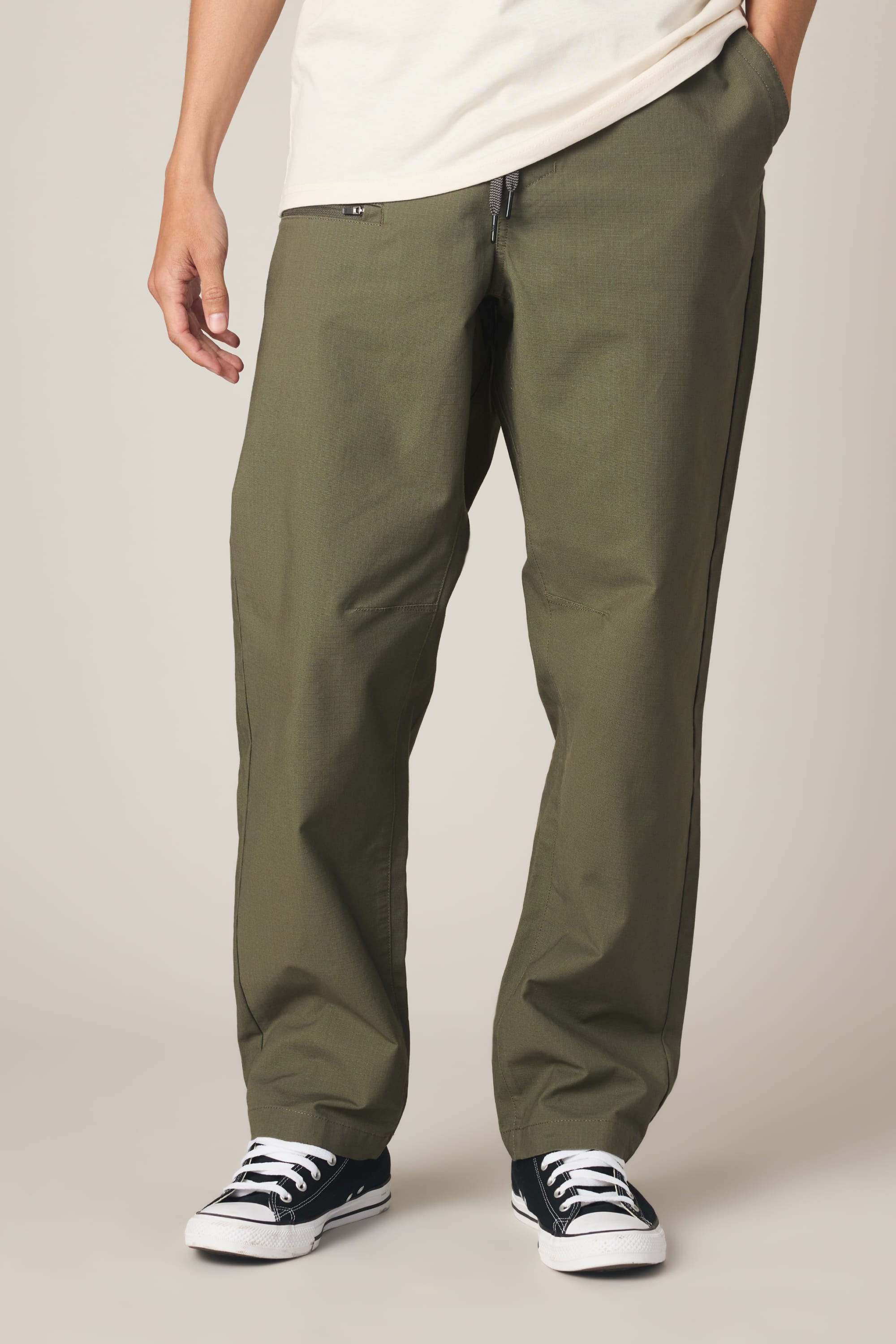 A person wears 686 Men's Free Loader Pant - Loose Fit in olive green, paired with a cream shirt and black low-top sneakers with white laces, standing on a light background with hands in pockets.