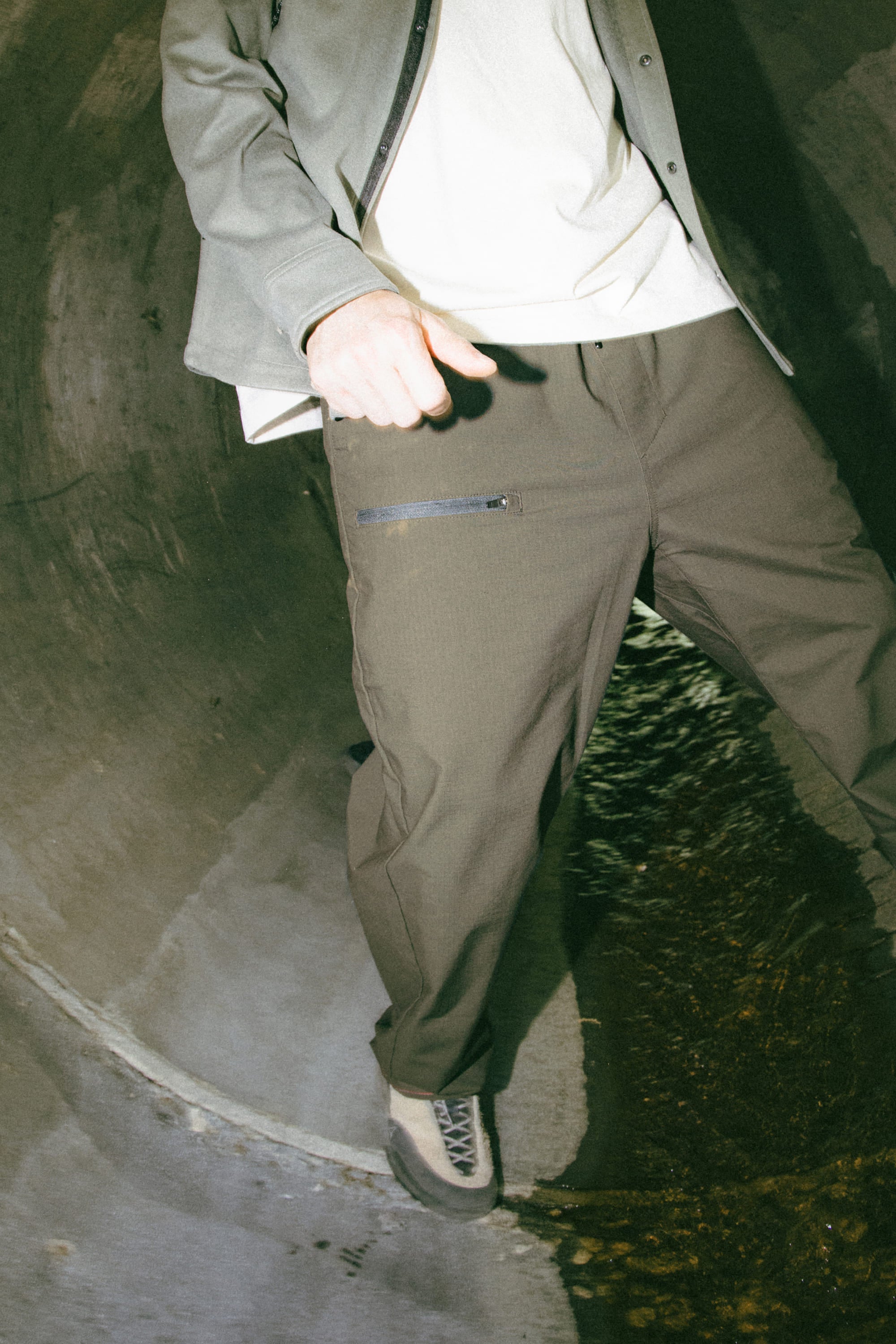A person stands in a dimly lit concrete tunnel, wearing a light shirt, jacket, and the 686 Men's Free Loader Pant - Loose Fit by 686, featuring a zipper pocket and crafted from durable nylon woven fabric, along with sneakers. Their face is not visible in the image.