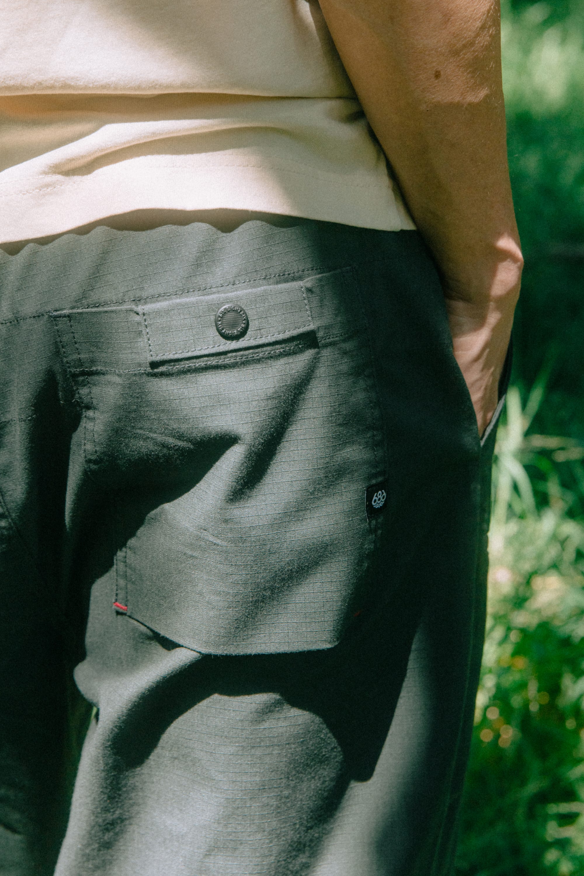 A person wearing the 686 Men's Free Loader Pant - Loose Fit in dark green and a beige shirt stands outdoors with one hand in their pocket.