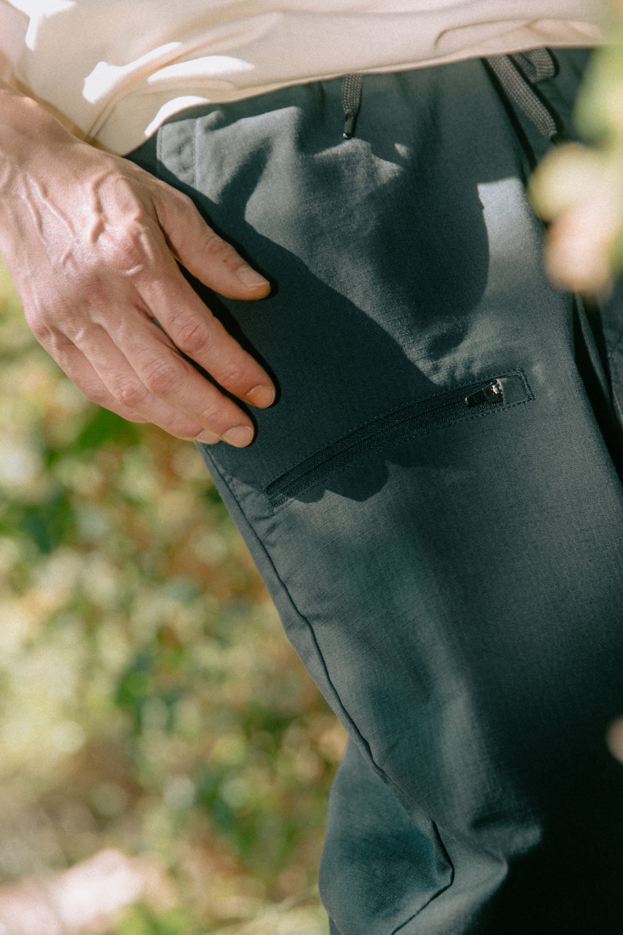 A close-up of a person's hand resting on the thigh of the dark-colored 686 Men's Free Loader Pant - Loose Fit, featuring a small zipper pocket, outdoors with greenery in the background. This pant from 686 is crafted from durable nylon, making it ideal for active days outside.