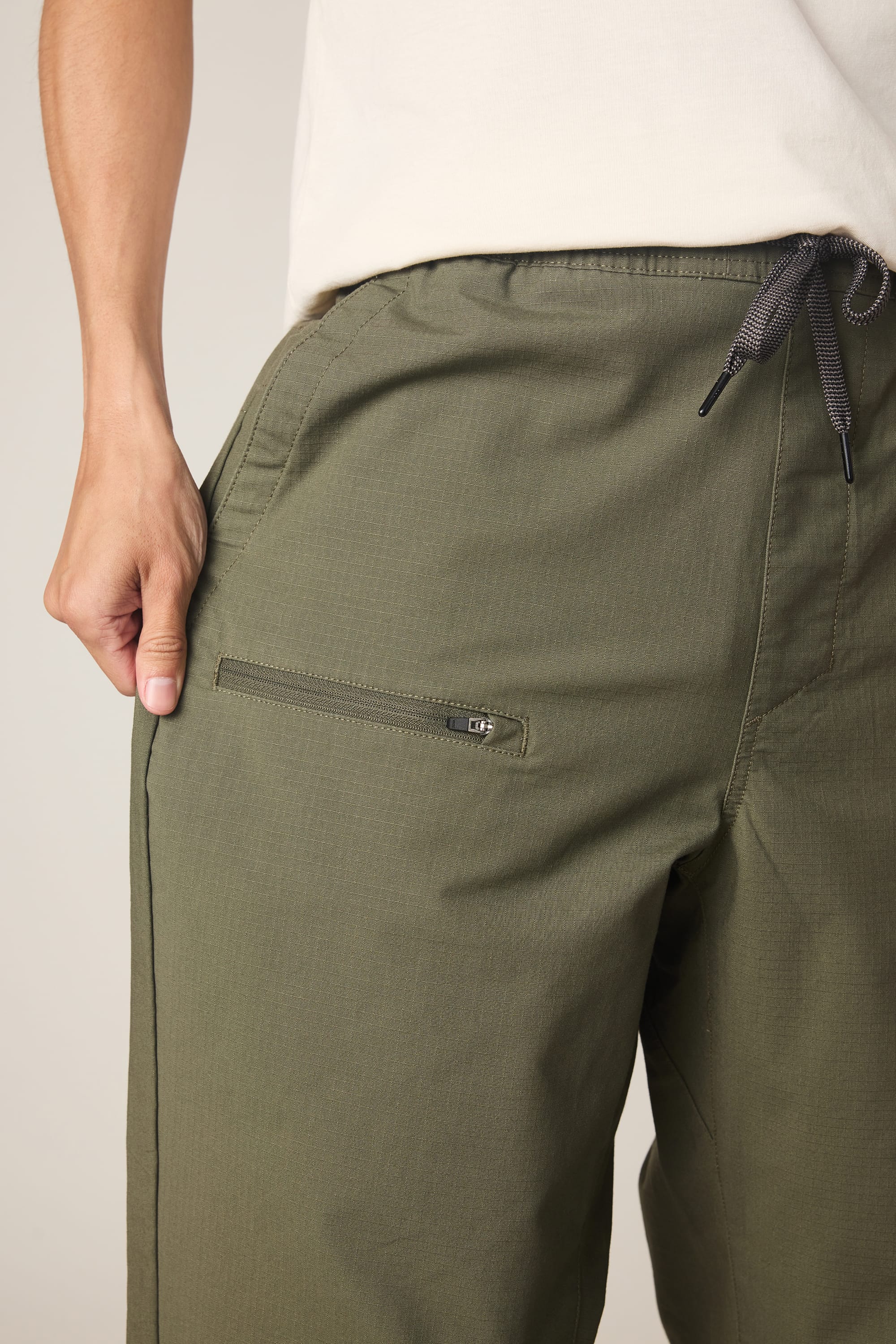Close-up of a person wearing the 686 Men's Free Loader Pant - Loose Fit in olive green, featuring a front zipper pocket and drawstring waistband. Made from nylon woven fabric by 686, paired with a white shirt, left hand in the pocket.