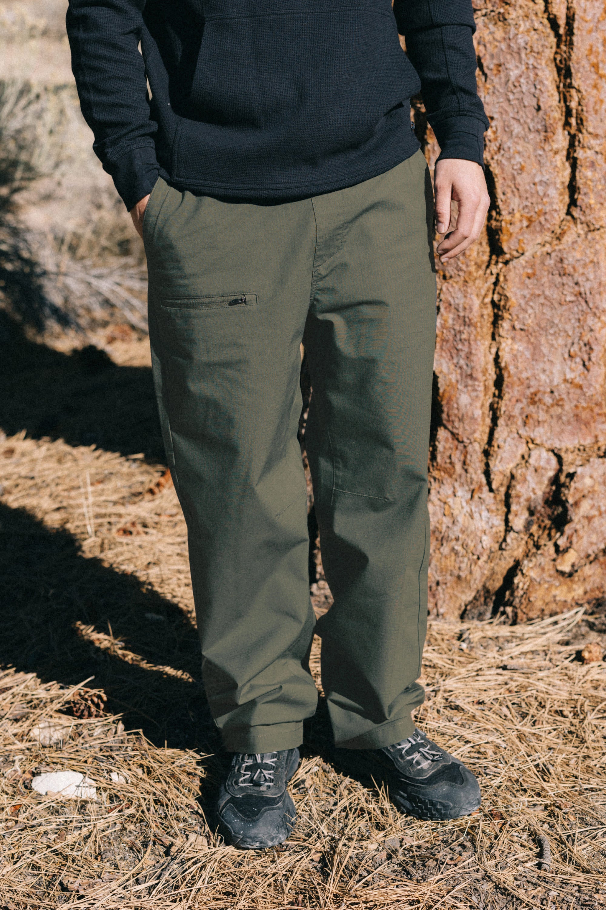 A person stands outdoors on dry grass near a large tree trunk, wearing a black sweatshirt, the 686 Men's Free Loader Pant - Loose Fit in green, and black shoes. Their face is not visible.