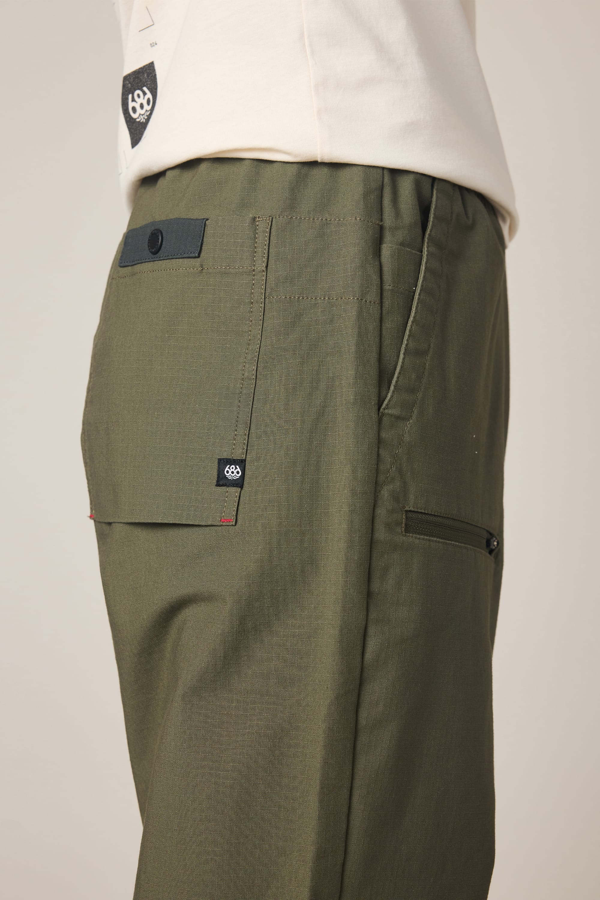A close-up side view of the 686 Men's Free Loader Pant - Loose Fit in olive green, featuring textured nylon fabric, a back pocket with black button, small logo tags near the pocket, and paired with a light-colored shirt.