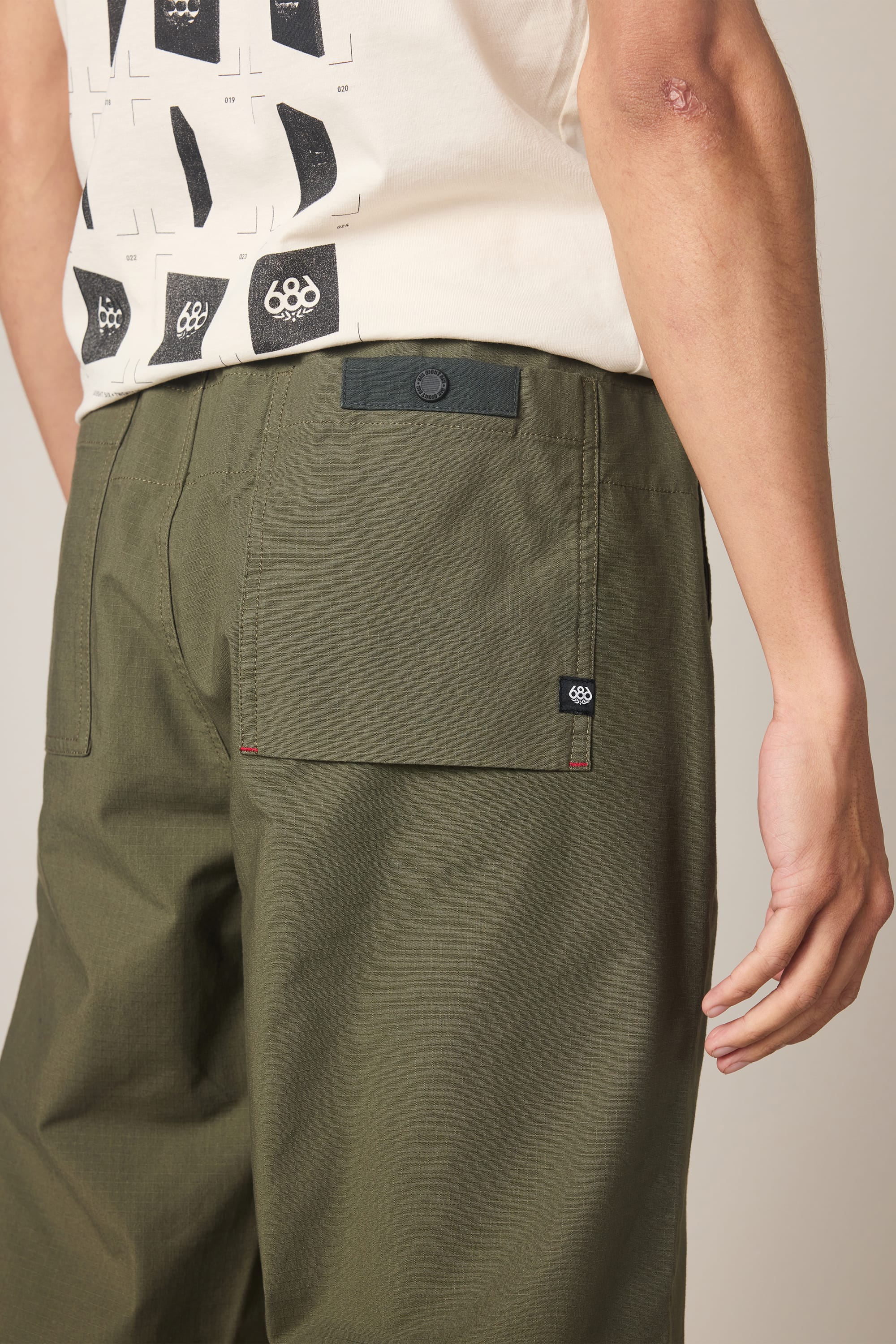 A person wearing 686 Men's Free Loader Pant - Loose Fit in olive green, featuring back pockets, paired with a white T-shirt with a black graphic. Their right arm shows a small mark near the elbow.