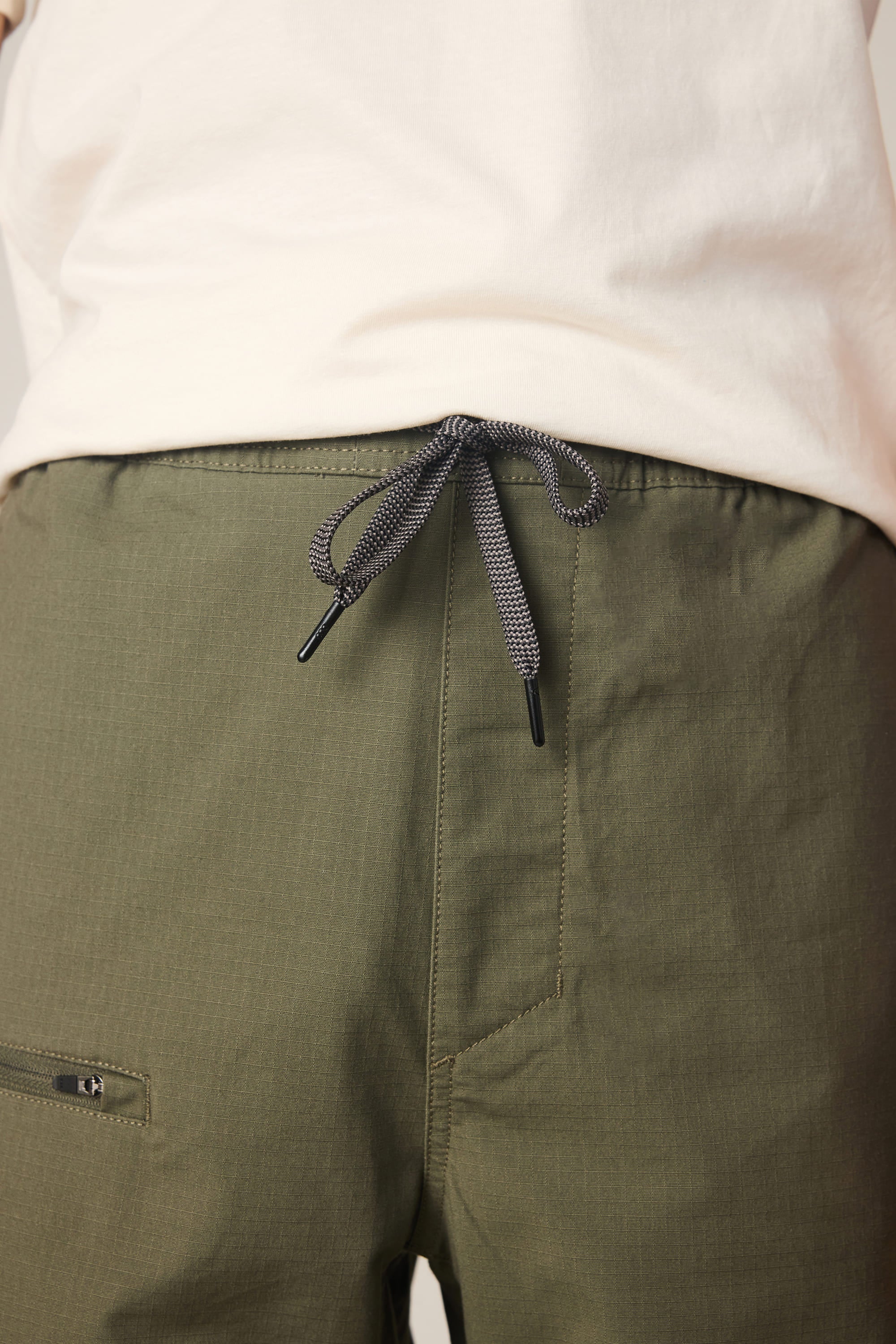 Close-up of a person wearing the 686 Men's Free Loader Pant - Loose Fit in olive green, featuring a black-and-white drawstring tied in a bow at the waistband. Paired with a white shirt, these comfortable spandex pants by 686 are finished with eye-catching detail at the waist.