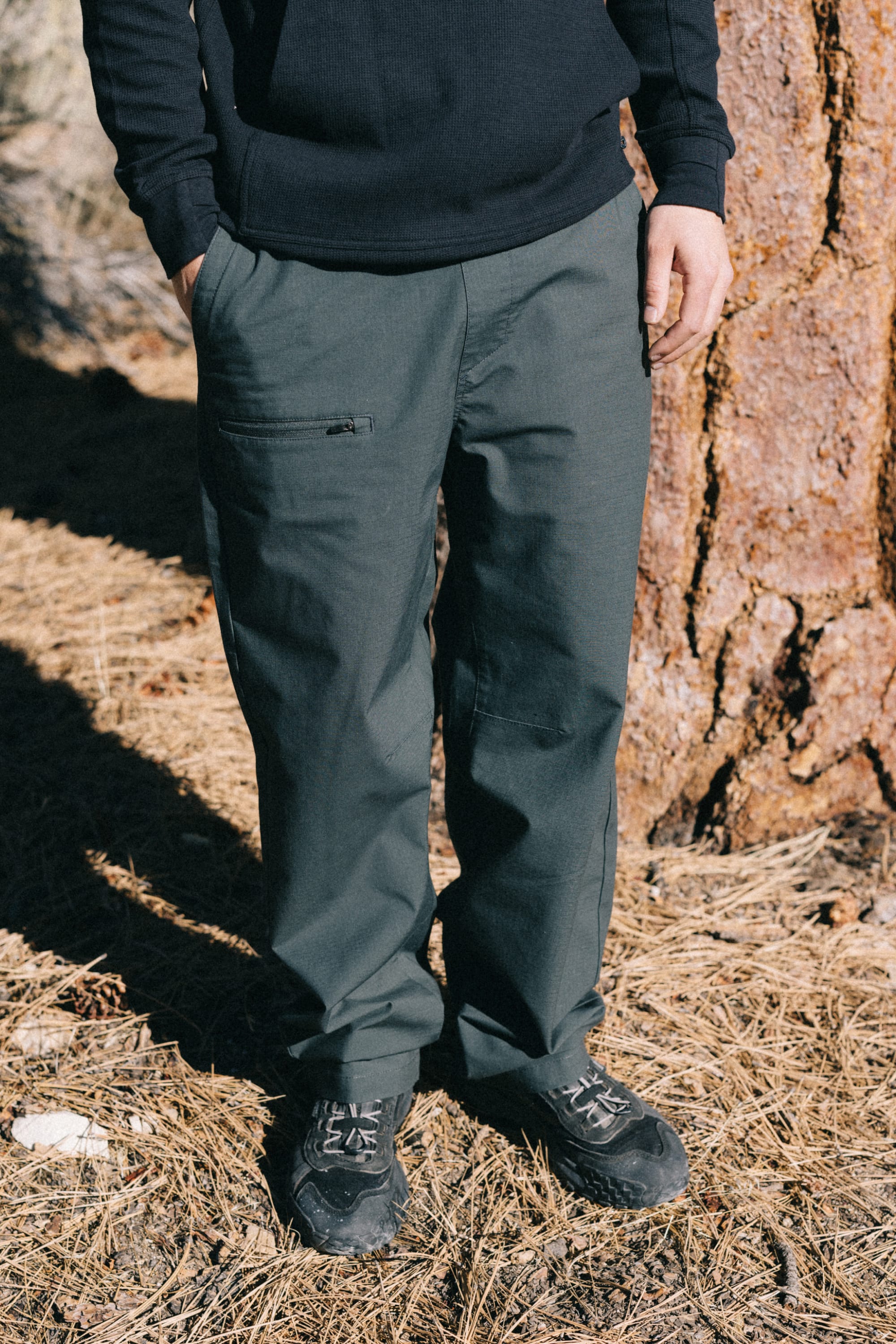 A person, whose face is not visible, stands outdoors on dry grass beside a tree wearing the 686 Men's Free Loader Pant - Loose Fit by 686 in dark green—a comfortable nylon woven men’s pant—paired with black shoes and a long-sleeve black shirt.