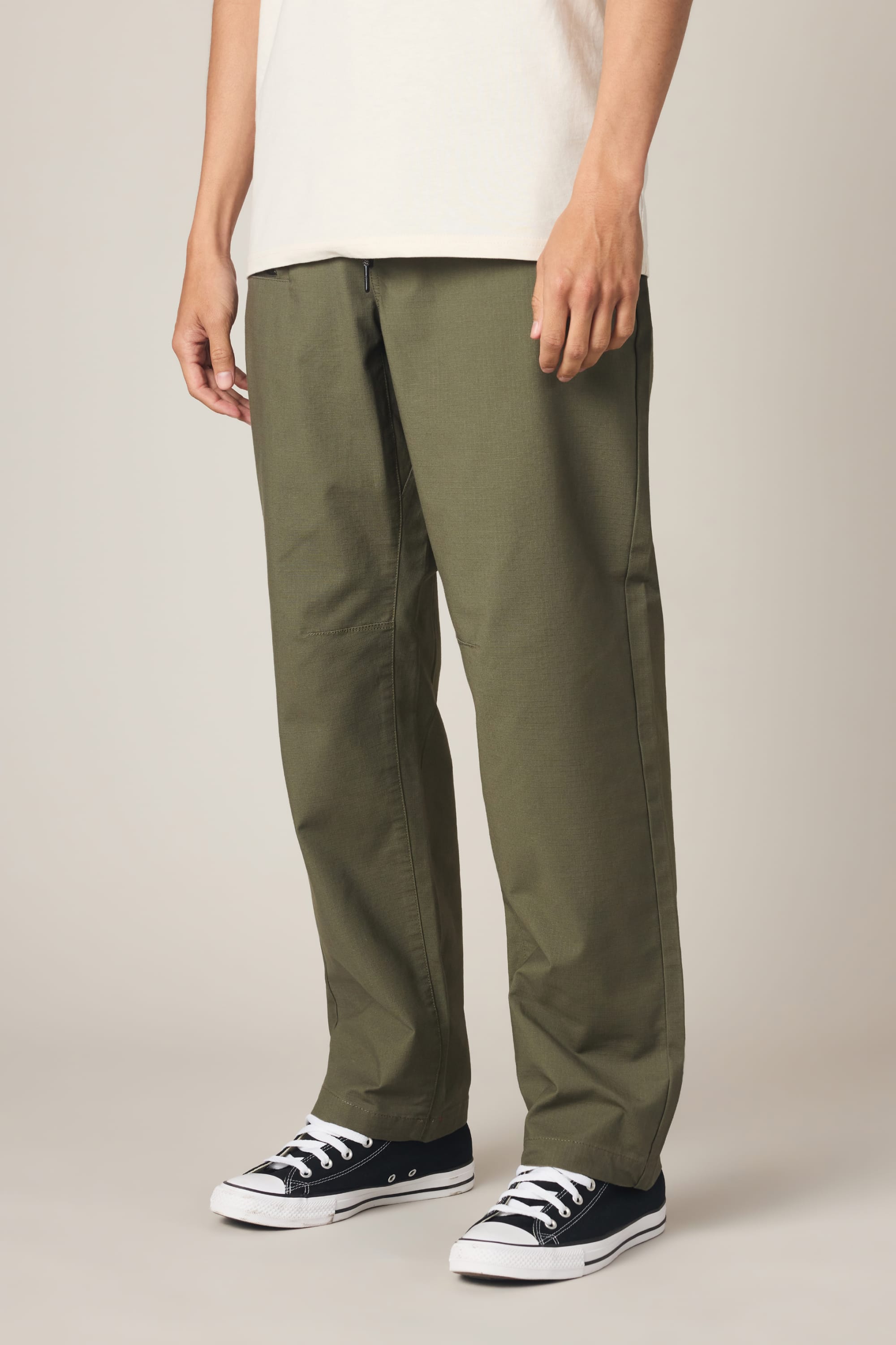 A person wearing 686 Men's Free Loader Pant - Loose Fit in olive green, a white T-shirt, and black low-top sneakers with white soles stands against a plain beige background.