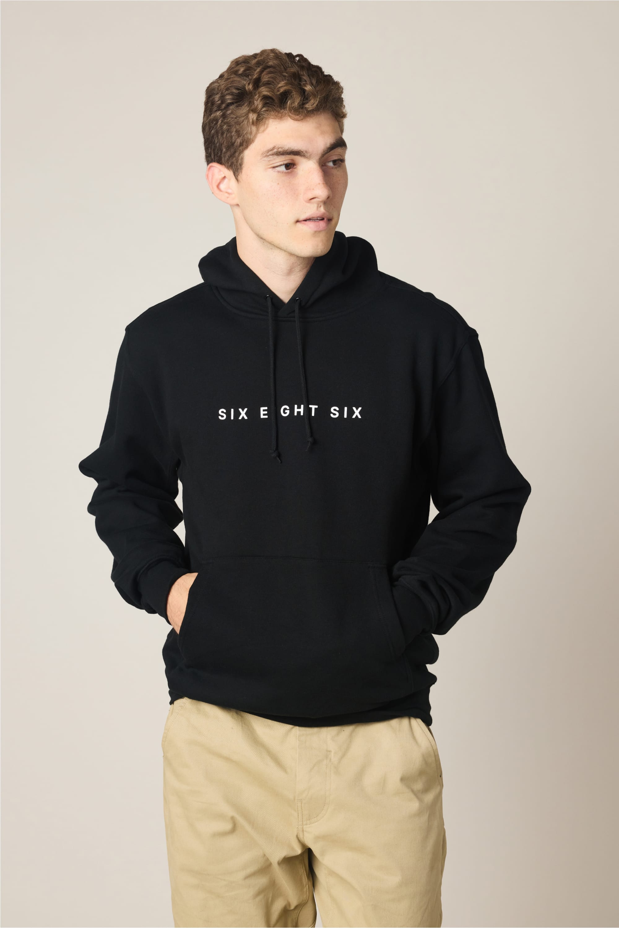 A young man in a 686 Wordmark Athletic LS Hoody‚Äîa black sweatshirt with ‚ÄúSIX EIGHT SIX‚Äù in white on the front‚Äîand light khaki pants stands with his hands in his pockets, looking off to the side against a plain background.