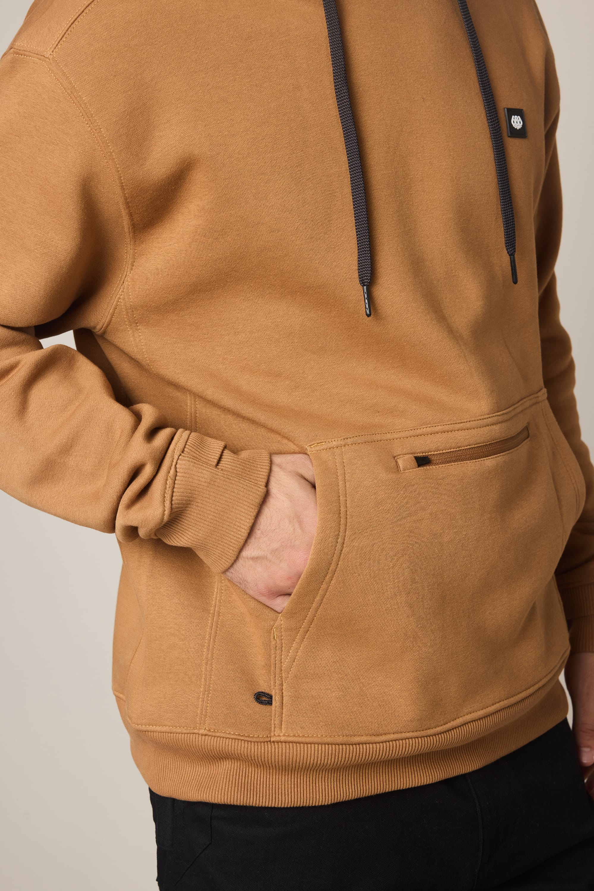 A person wears the 686 Men's All Season Hoody—a tan cotton sweatshirt with dark drawstrings, a front kangaroo pocket, and a small black chest logo patch—paired with black pants.