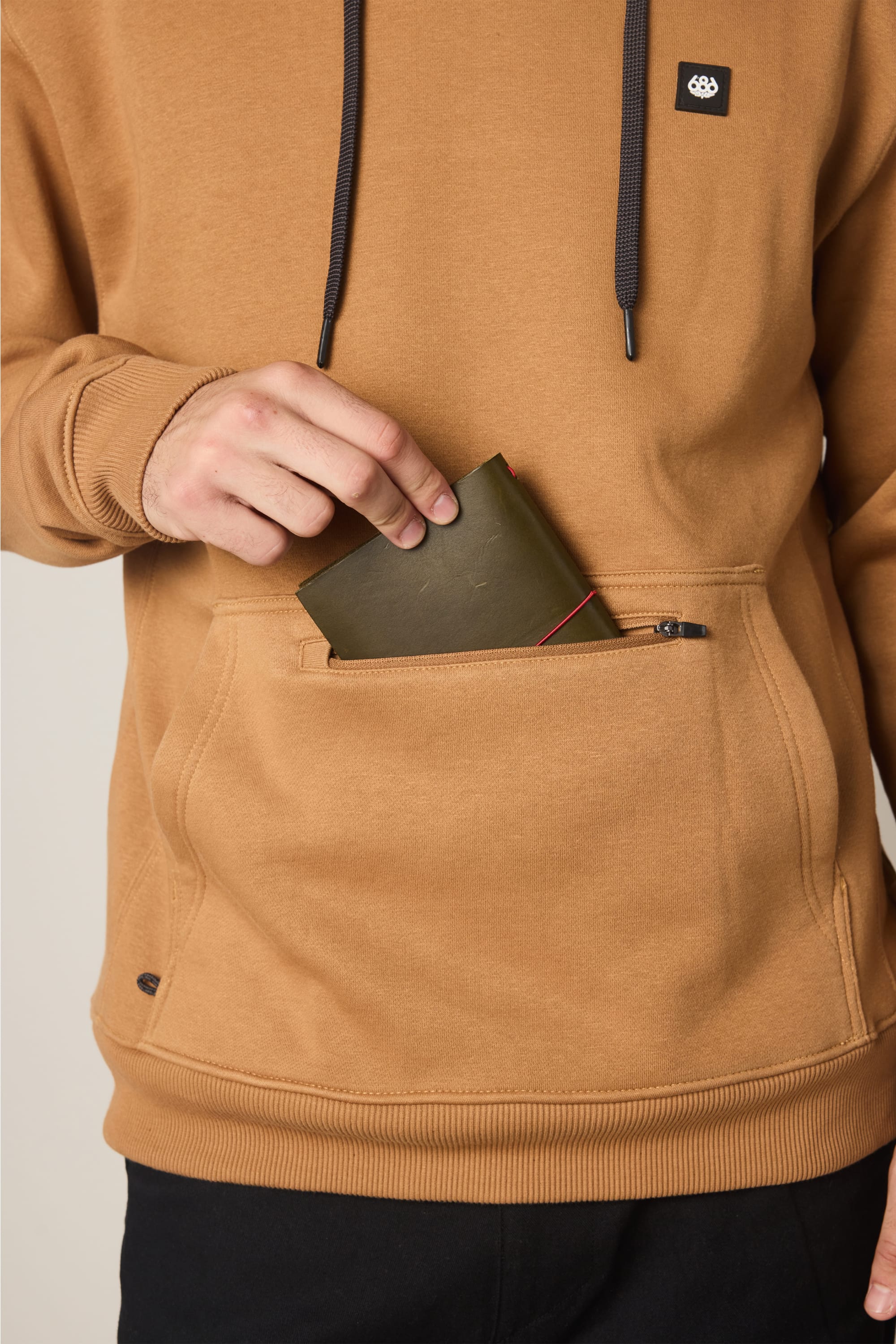 A person wearing a 686 Men’s All Season Hoody puts a green wallet with red edges into the front zippered pocket of the sweatshirt.