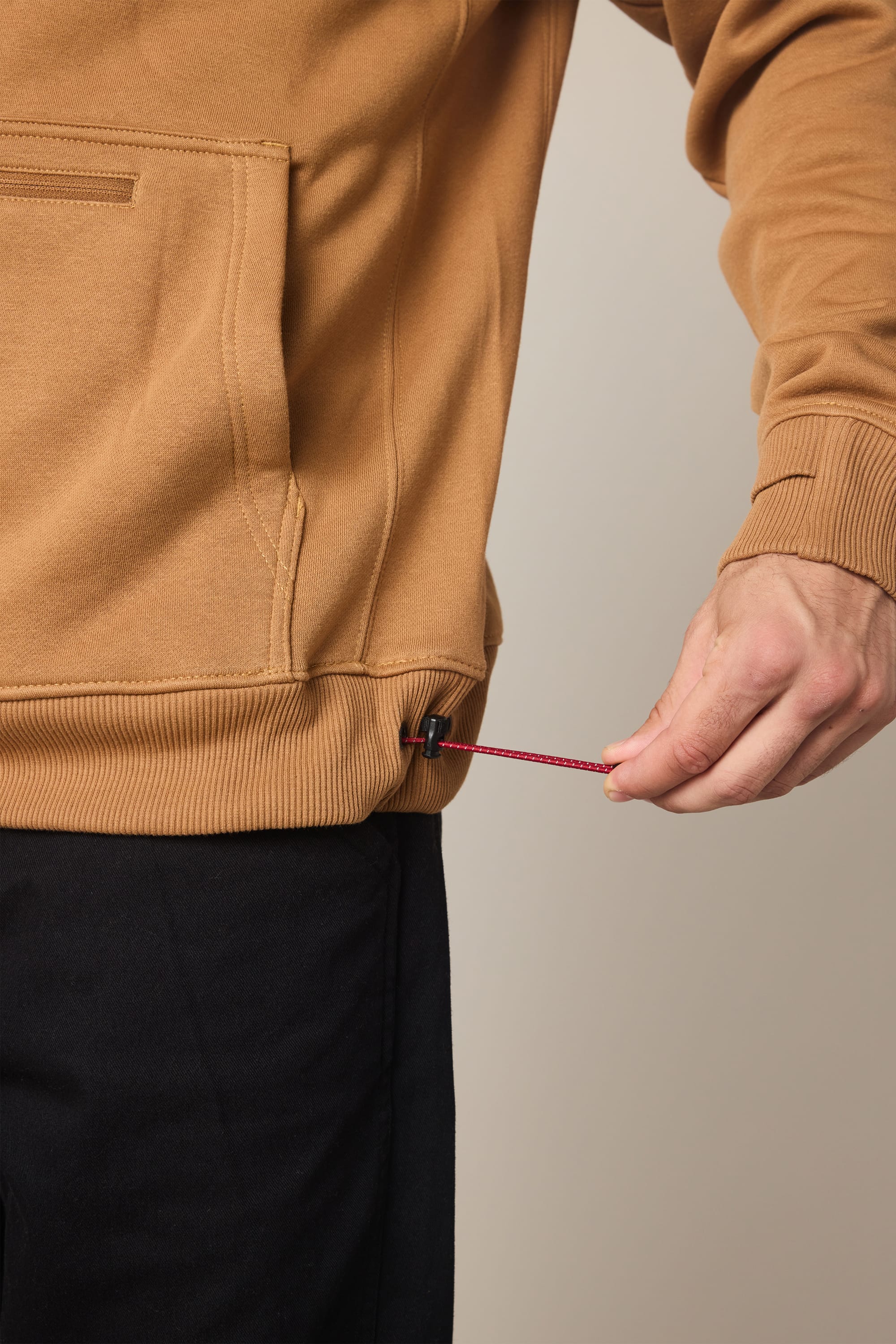 A person is adjusting the drawstring at the hem of a tan 686 Men's All Season Hoody, paired with black pants. Only their torso and right hand are visible.