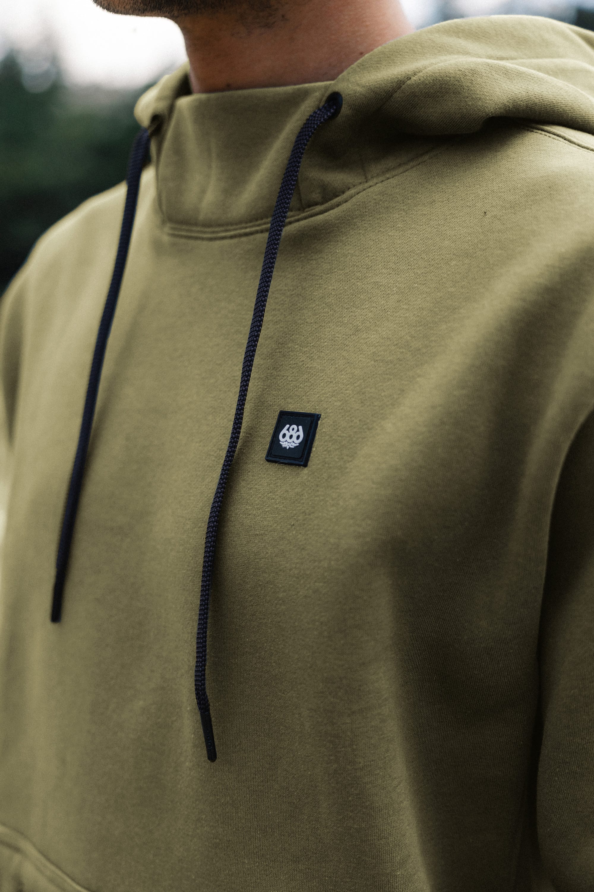 A person wearing the 686 Men's All Season Hoody in olive green, featuring black drawstrings and a small black embroidered 686 logo patch on the chest. The cotton sweatshirt is shown in a photo cropped to display only the upper torso.