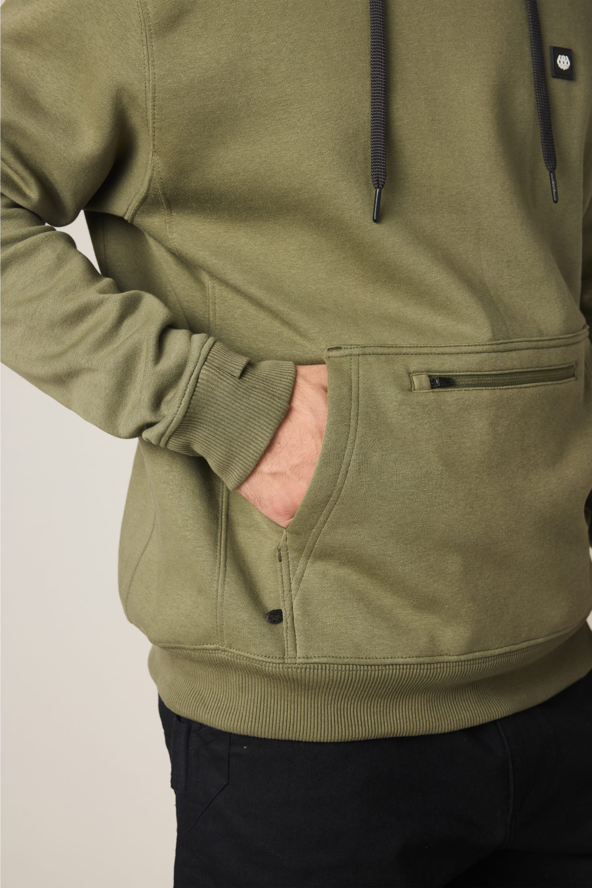 A person wearing the 686 Men's All Season Hoody—an olive green cotton sweatshirt by 686 featuring a front pocket and black drawstrings—stands with one hand in the pocket. Only the torso and part of the upper legs, dressed in black pants, are visible. This comfortable and stylish hoody is perfect for all seasons.