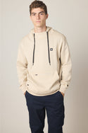 A young man with short curly hair wears the 686 Men's All Season Hoody in beige with black drawstrings and a small logo patch, paired with dark navy pants, standing hands-in-pockets against a plain background.