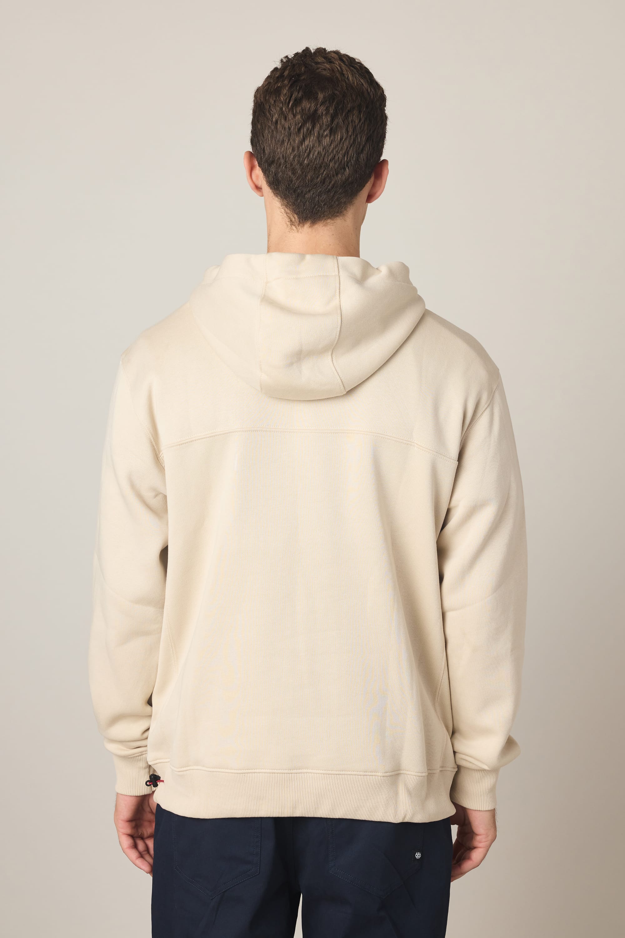 A person with short brown hair stands with their back to the camera, wearing a light beige 686 Men's All Season Hoody and dark blue pants against a plain light background.