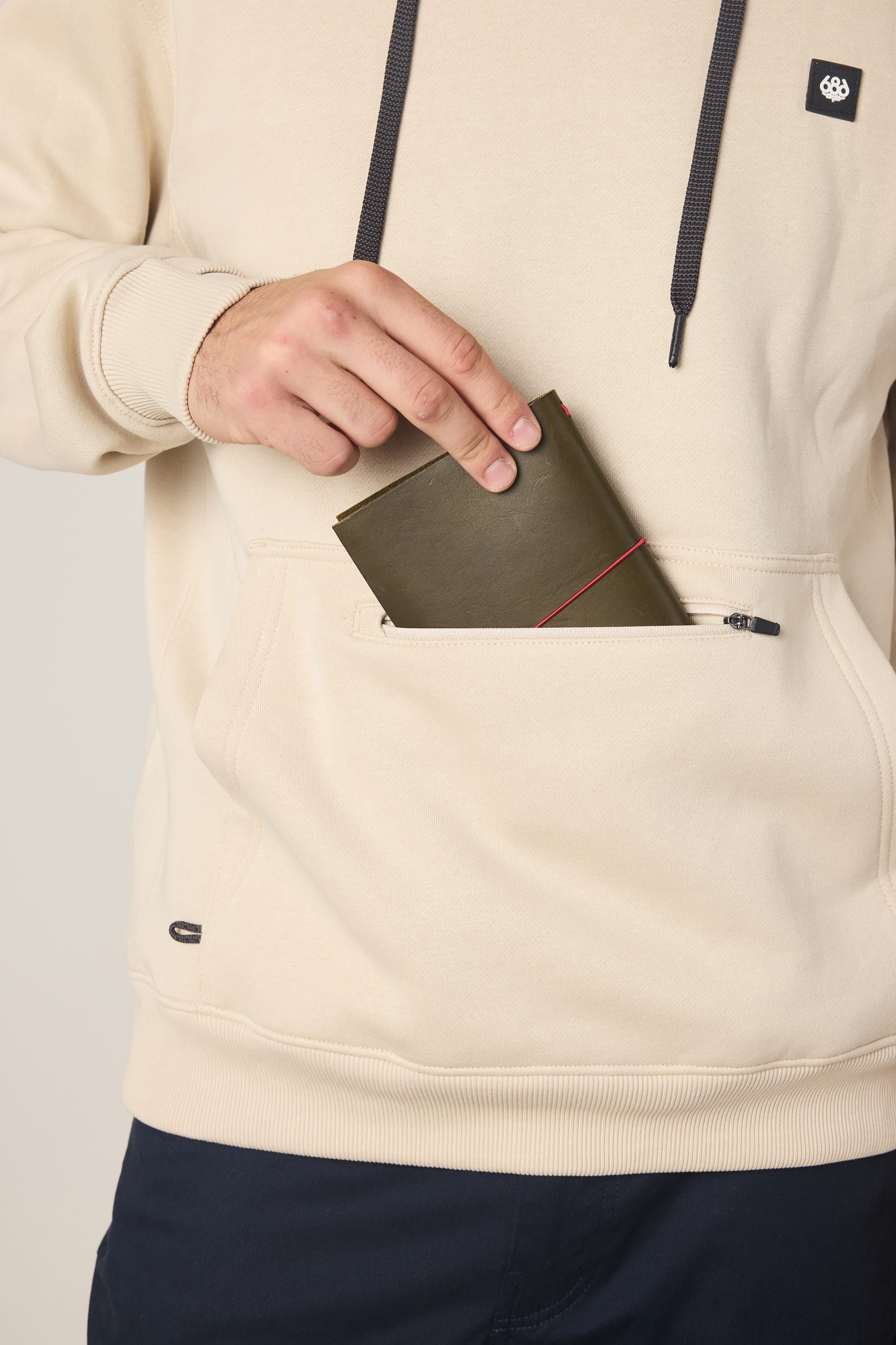A man in a beige 686 Men's All Season Hoody and dark pants puts a green wallet with a red accent into the front zippered pocket of his cotton sweatshirt.