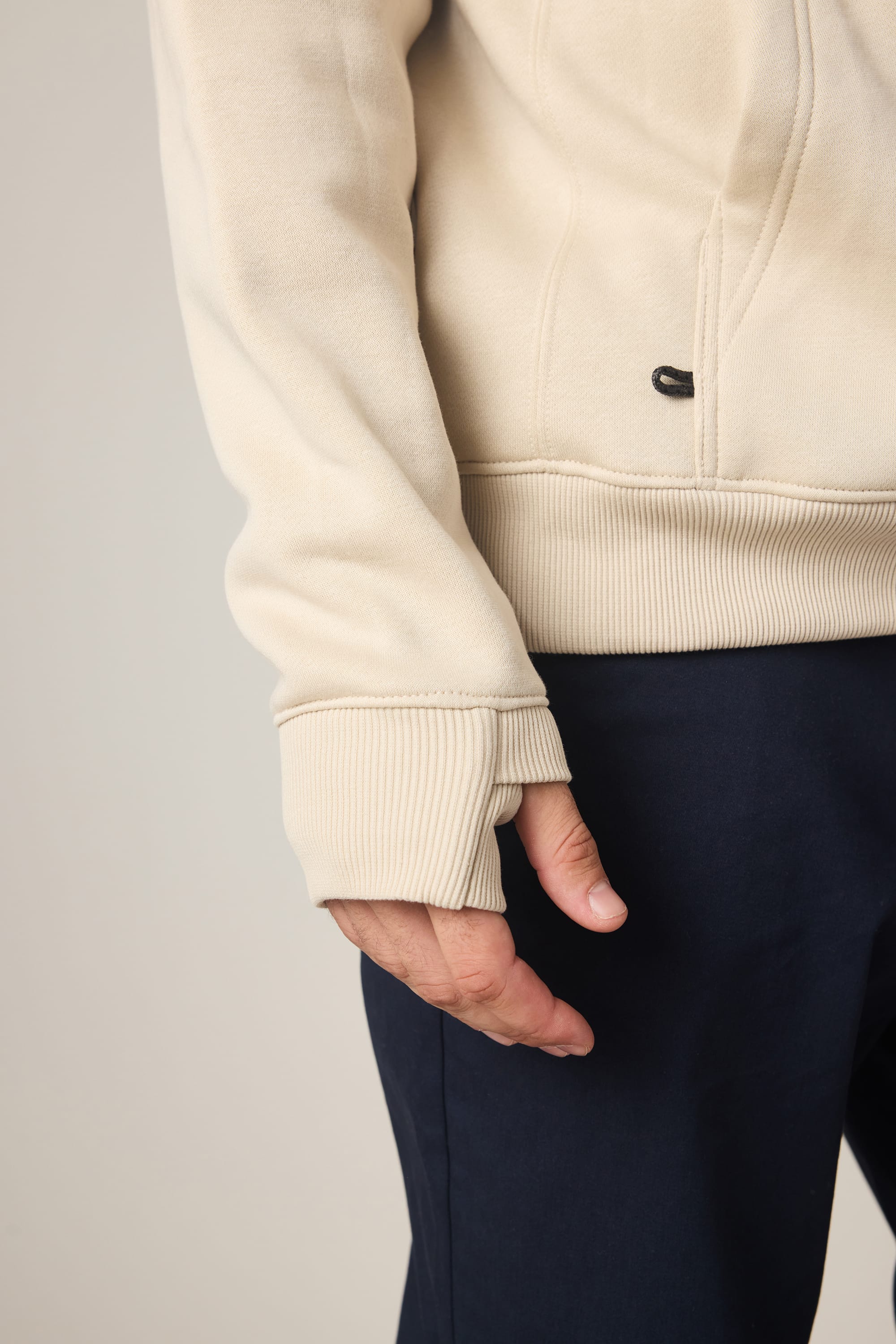 A person wearing the 686 Men's All Season Hoody in cream and dark pants stands with their left arm relaxed by their side, highlighting the hoody’s ribbed cuff and thumbhole detail.