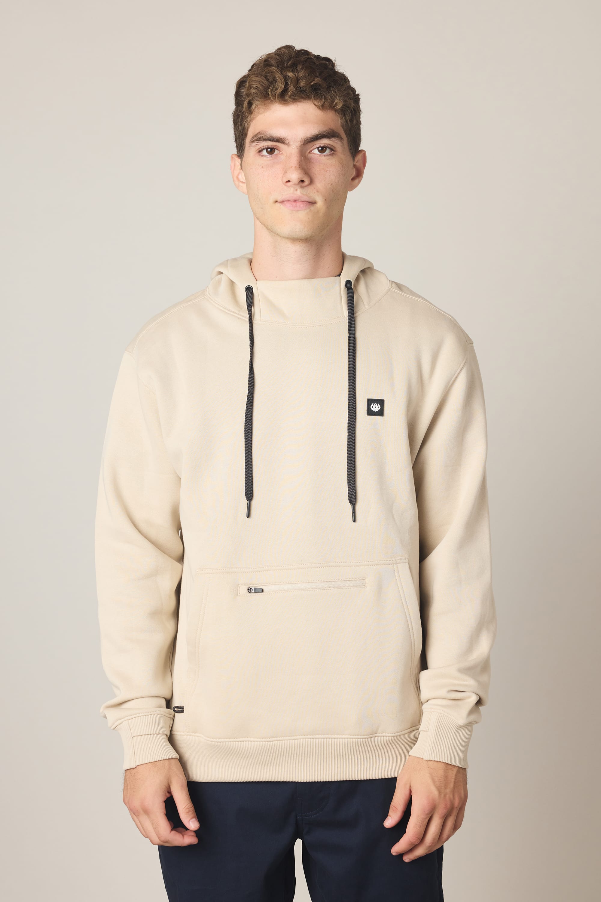 A young man with short curly hair wears the 686 Men's All Season Hoody, a beige cotton sweatshirt with black drawstrings and a small chest logo from 686, standing against a plain light background with a neutral expression.