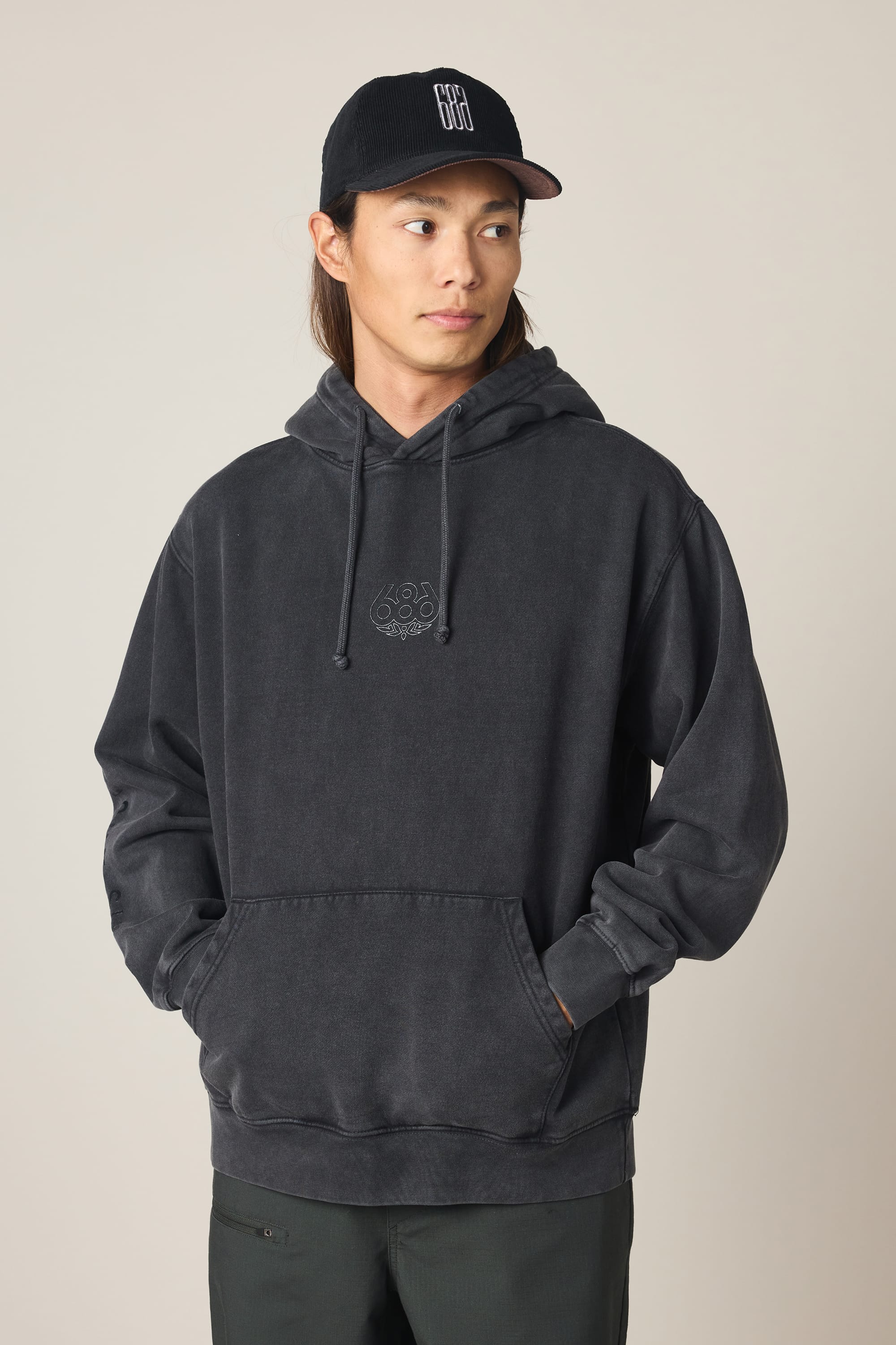 A man in the 686 Men's High Def Relaxed LS Hoody, featuring a small chest logo, black pants, and a black baseball cap stands with hands in the hoodie pocket, looking slightly to the side against a plain background.