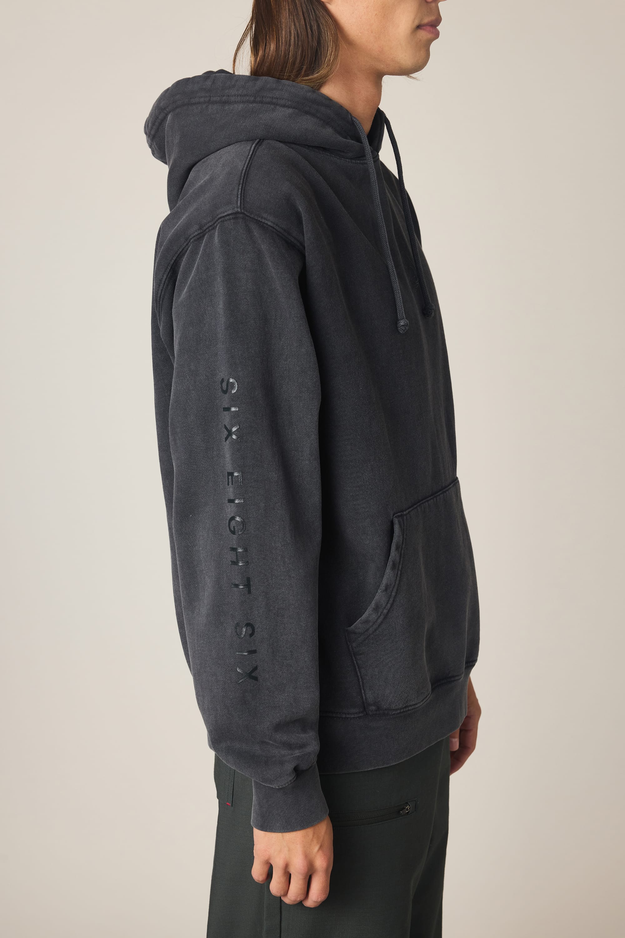 A person with long hair wears the 686 Men's High Def Relaxed LS Hoody, a black cotton blend sweatshirt featuring "SIX EIGHT SIX" printed vertically on the sleeve, standing sideways against a plain background.