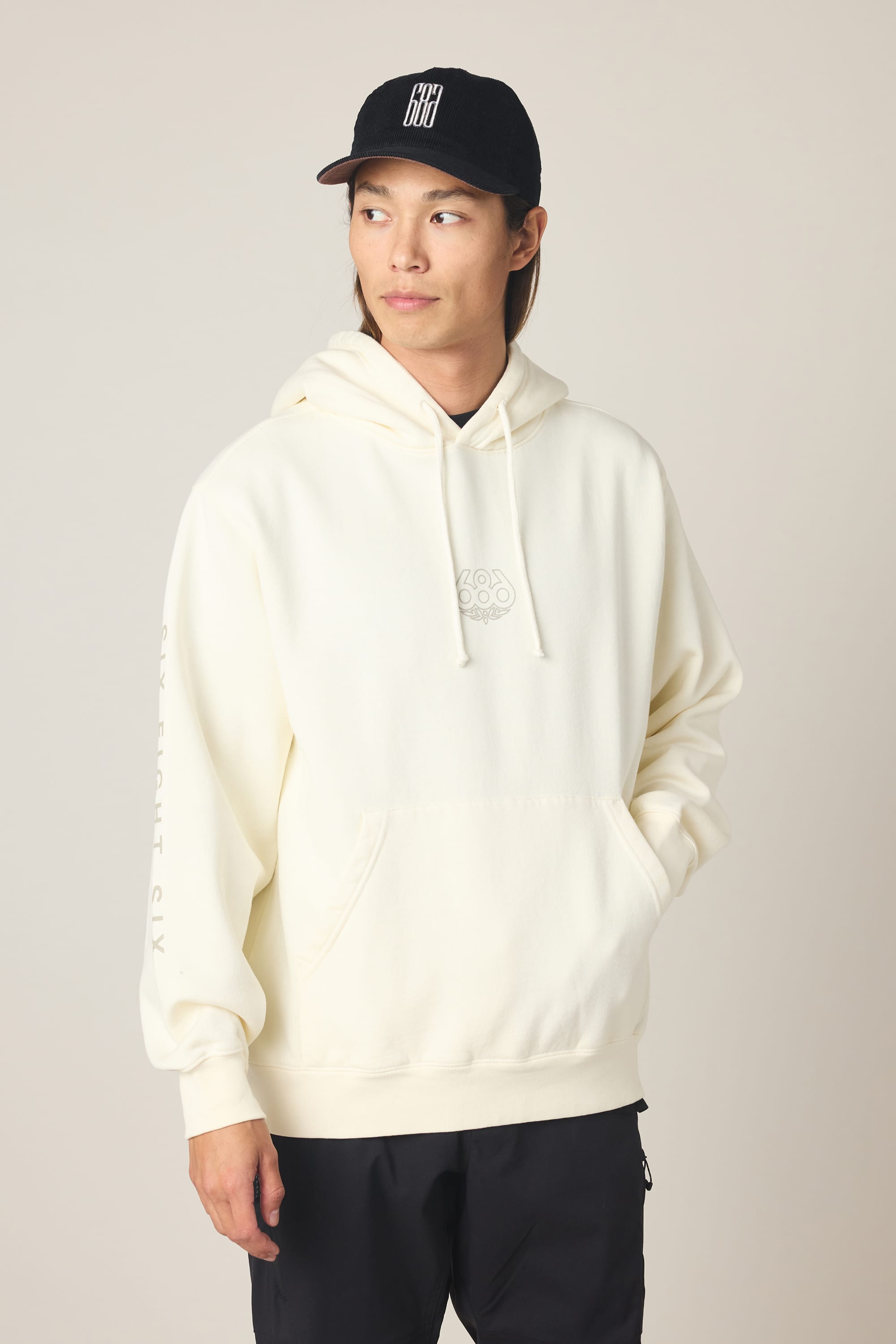 A person in a black baseball cap, black pants, and the 686 Men's High Def Relaxed LS Hoody—a relaxed off-white cotton sweatshirt with front pocket and subtle logo—stands with one hand in their pocket against a neutral background.