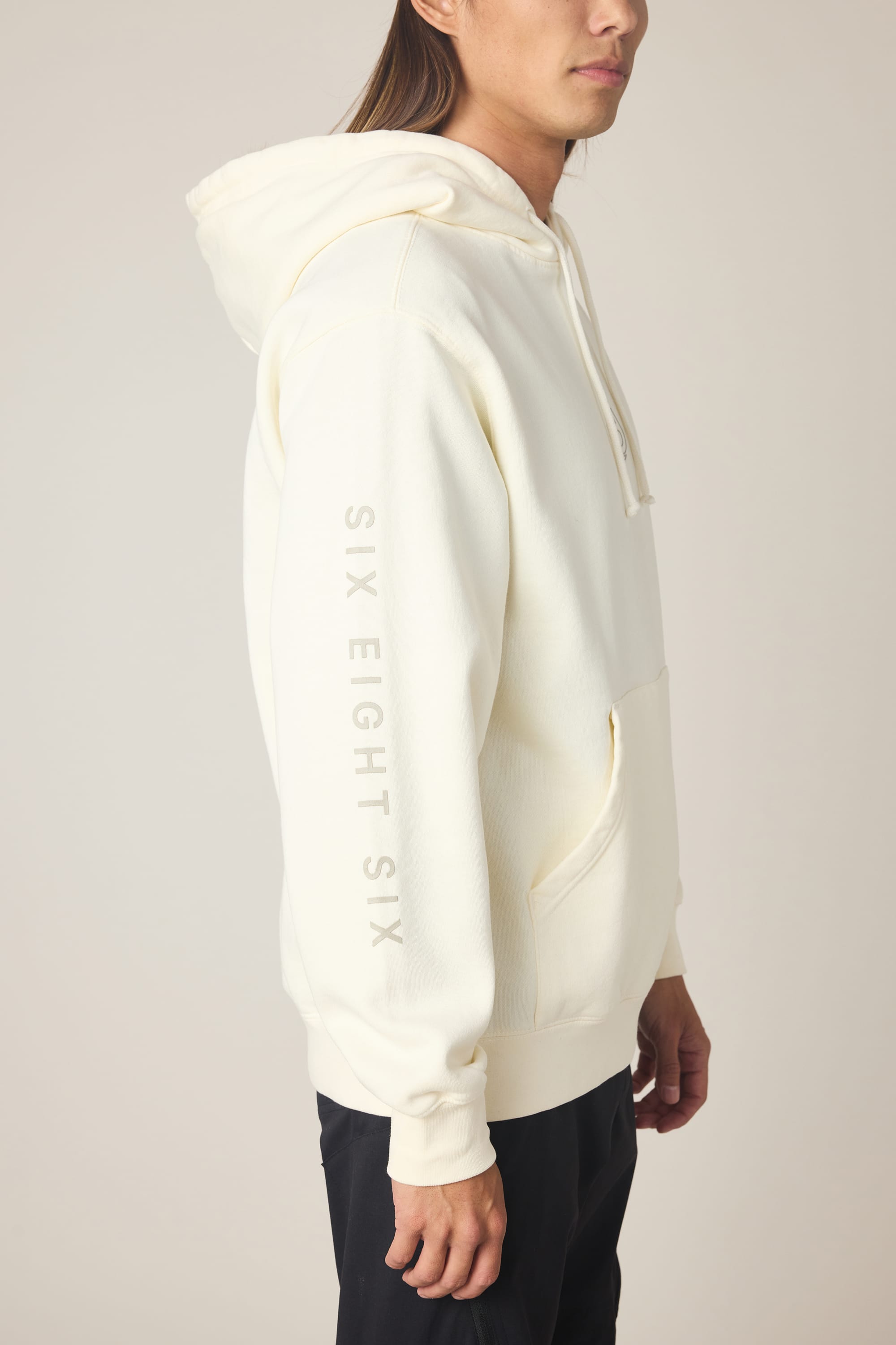A man stands sideways against a plain background, wearing the 686 Men's High Def Relaxed LS Hoody—an off-white cotton blend hoodie with a front pocket and "SIX EIGHT SIX" printed vertically down the sleeve.