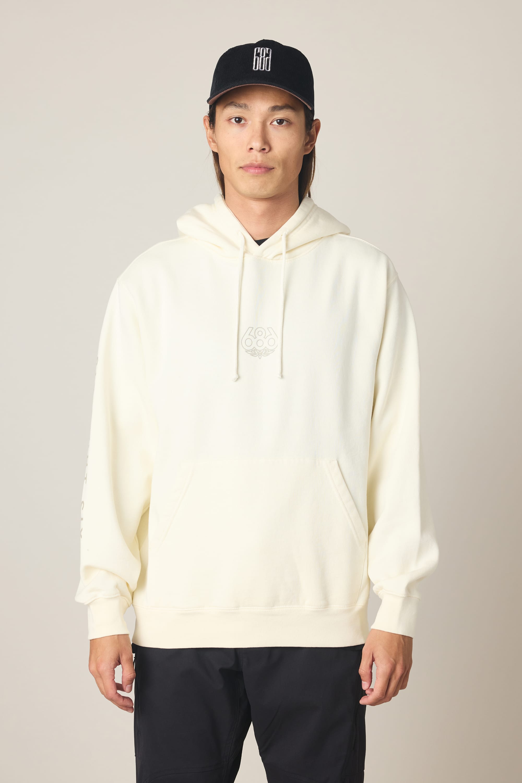 A person wearing the 686 Men's High Def Relaxed LS Hoody in cream with a subtle front logo and a black cap, standing against a plain light background.