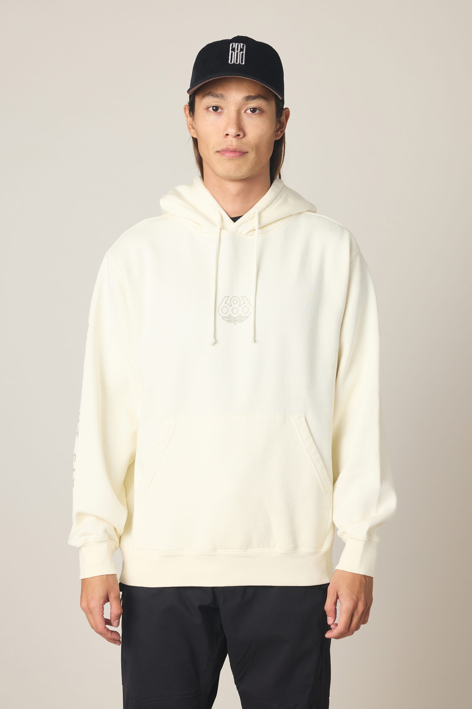 A person wearing the 686 Men's High Def Relaxed LS Hoody in cream, featuring a subtle chest logo, and a black baseball cap stands facing forward against a plain light background.