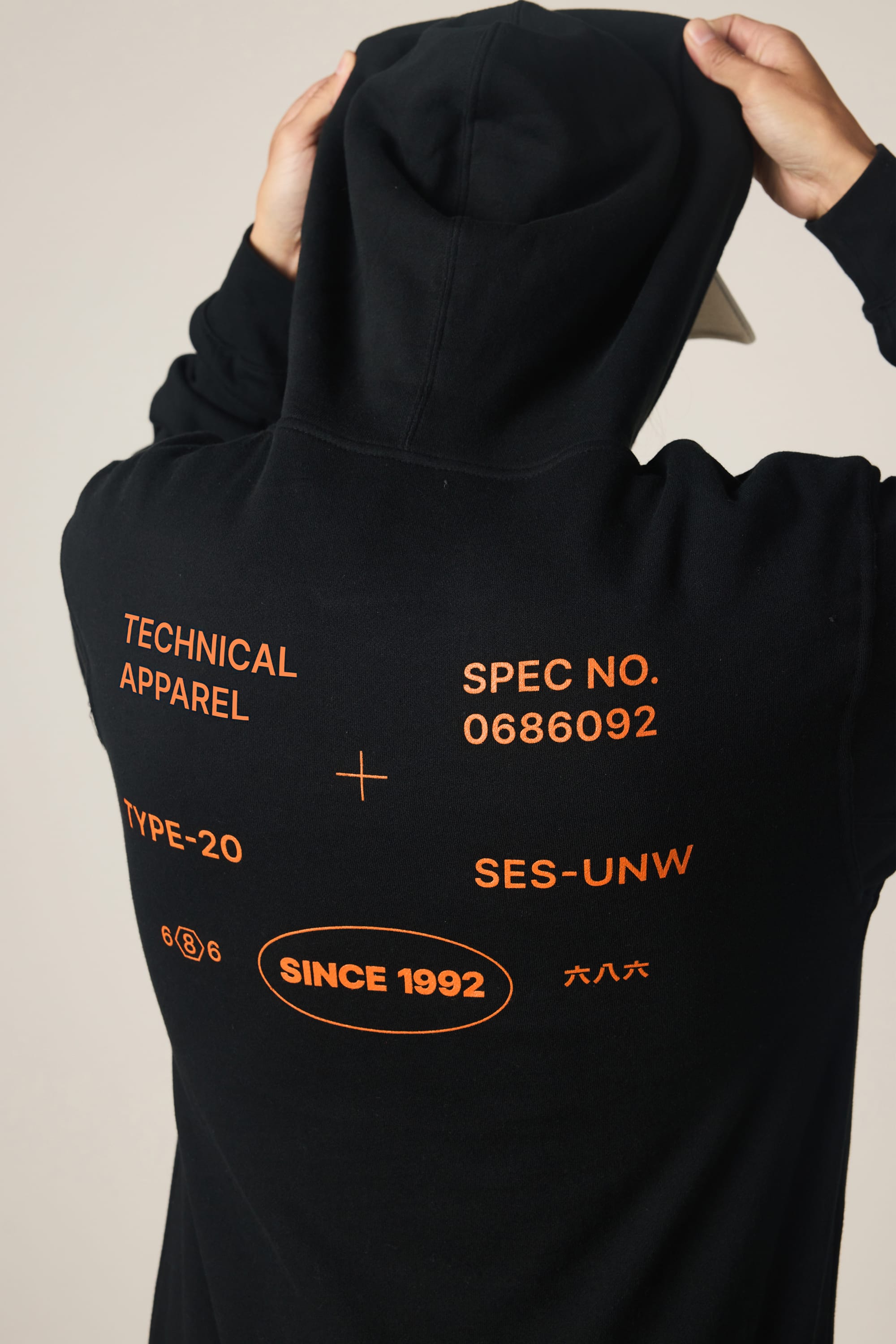 A person wears the 686 Men's Unwork Spec Athletic LS Hoody, a black cotton sweatshirt by 686, featuring orange text and graphics on the back with phrases like TECHNICAL APPAREL, SPEC NO. 0686092, TYPE-20, SES-UNW, and SINCE 1992 in an oval.