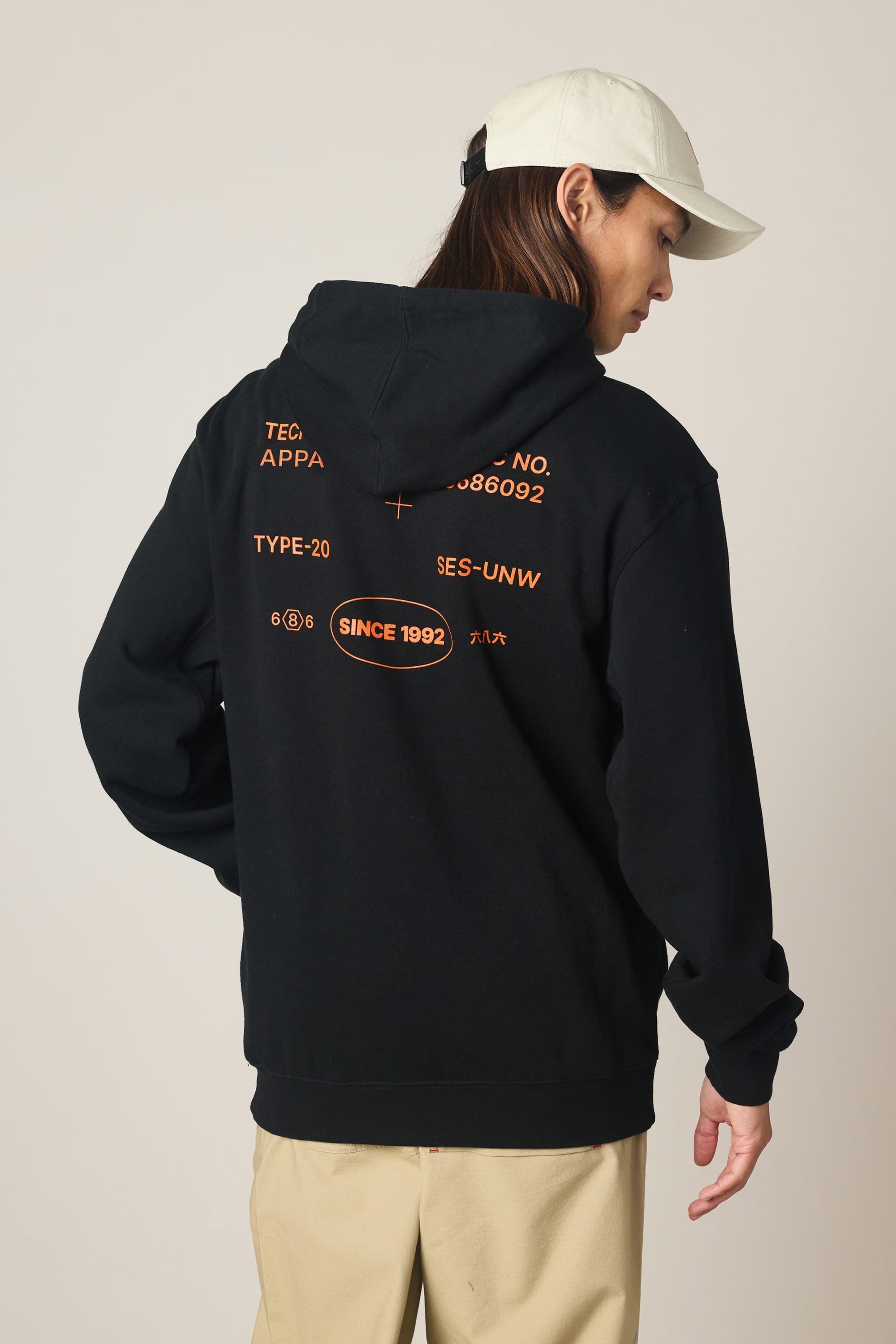 A person in a white cap and 686 Men's Unwork Spec Athletic LS Hoody stands facing away, showcasing the black sweatshirt’s orange text and graphics on the back. Beige pants complement the cotton hoody against a plain, light-colored background.