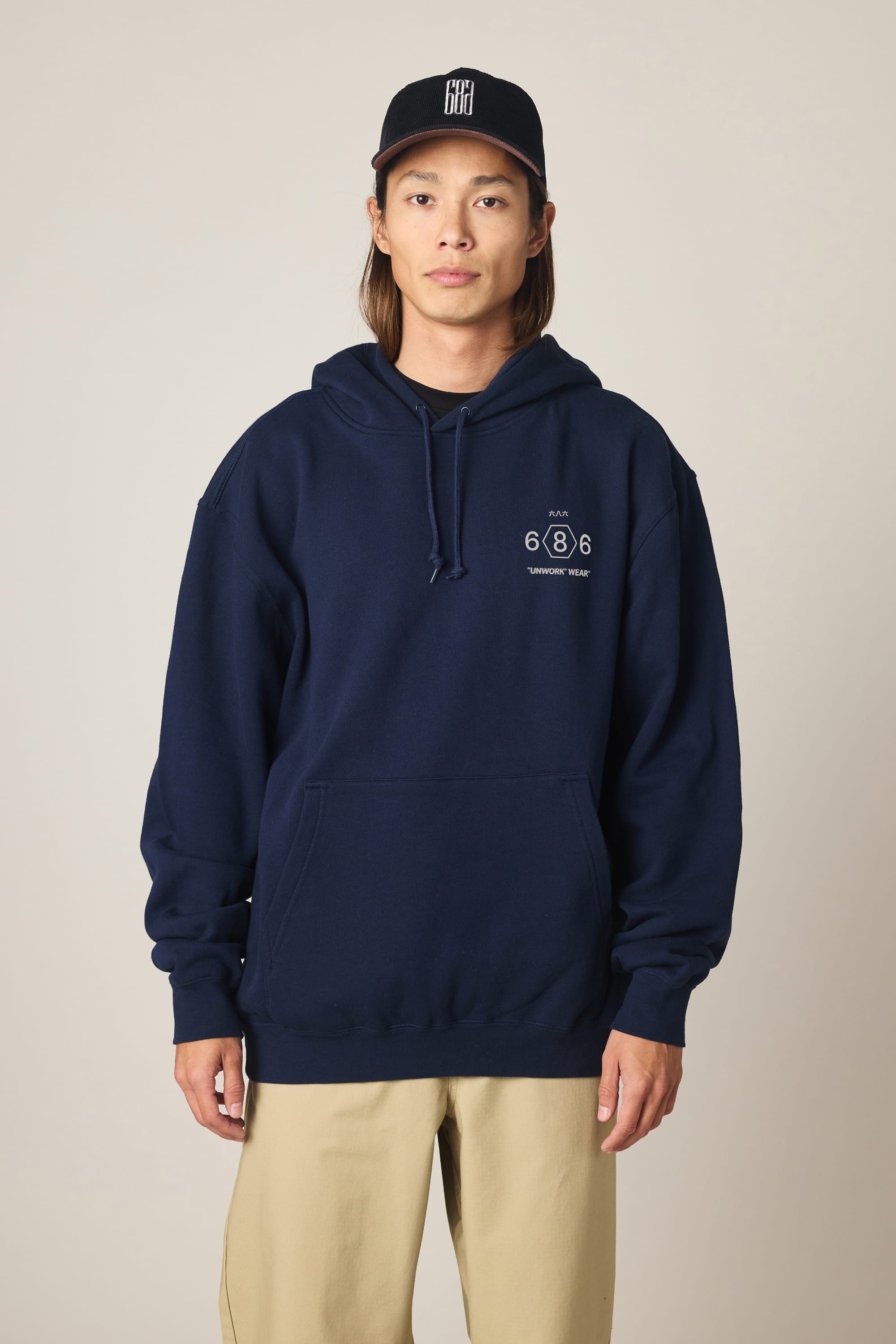 A person wearing the 686 Men's Unwork Spec Athletic LS Hoody in navy, featuring the 686 logo and text on the left chest, paired with beige pants and a black baseball cap, stands against a plain beige background.