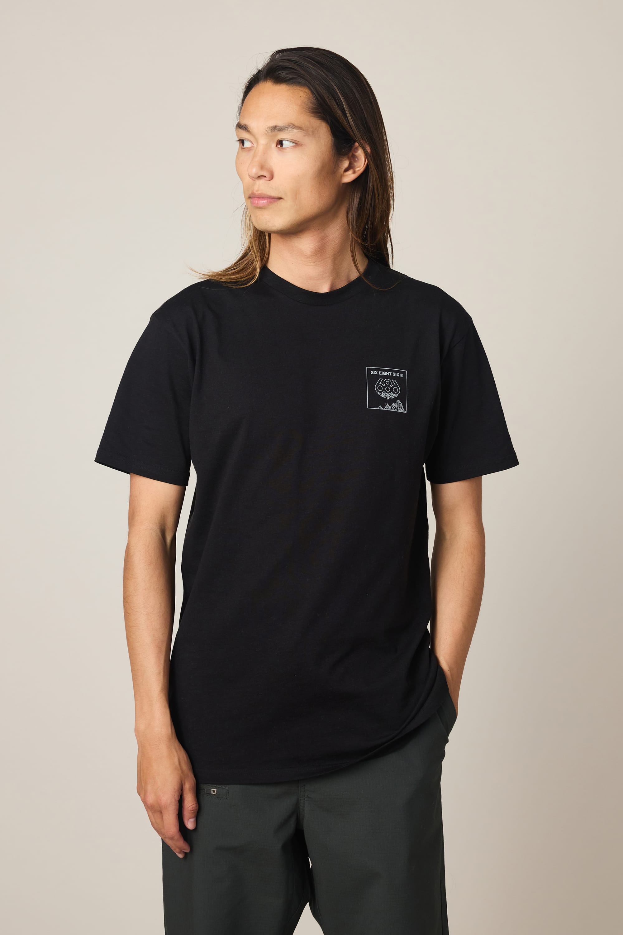 A person with long hair wearing a black 686 Cross-Cut Authentic S/S Tee by 686 and dark pants stands with one hand in their pocket, looking to the side against a plain light background.