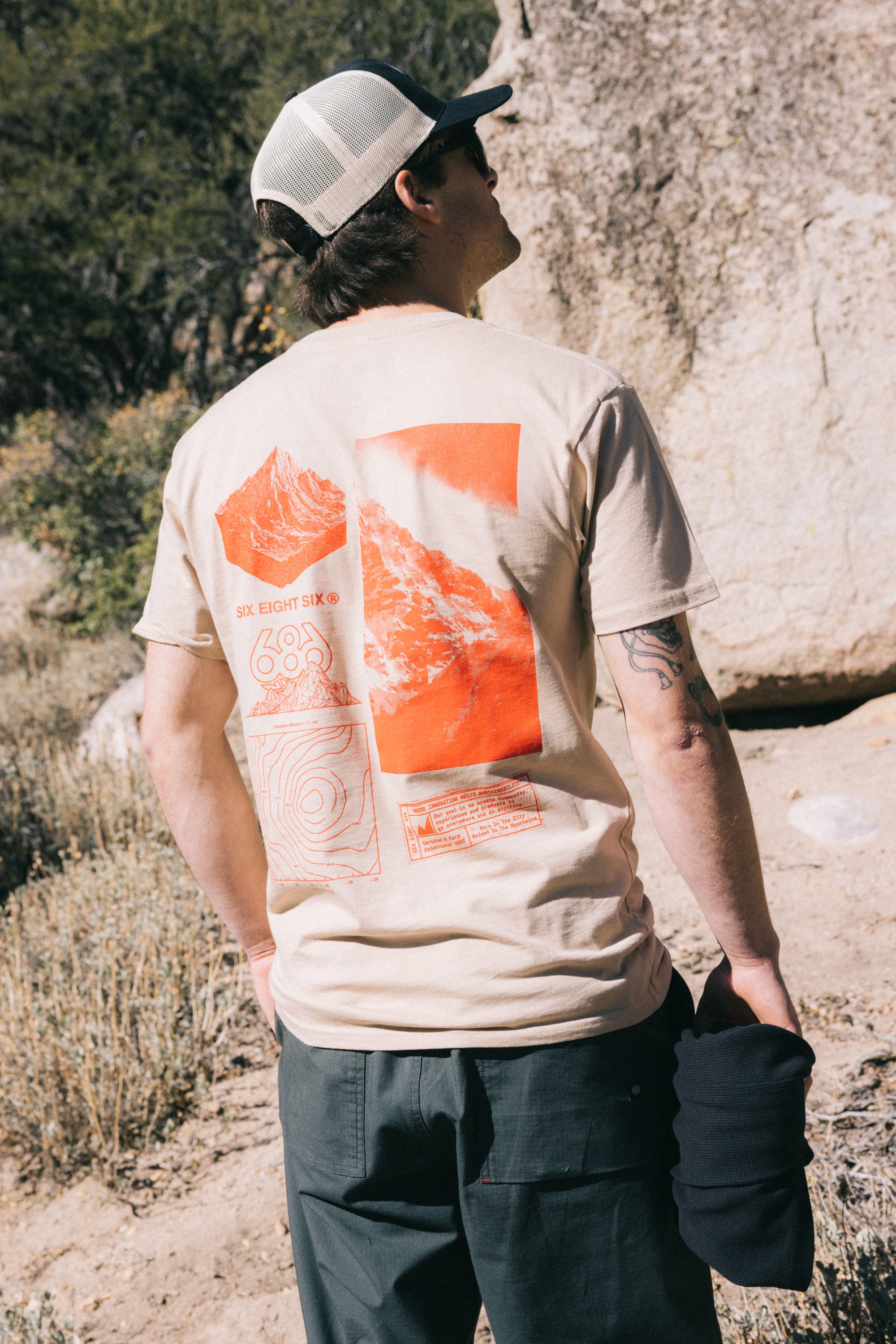 A person outdoors wears the 686 Cross-Cut Authentic S/S Tee, a beige men's t-shirt from 686 featuring bold red graphics of a mountain and topographic map on the back. Crafted from 100% cotton for comfort and style, the tee is paired with dark pants and a cap as the wearer stands near rocks and dry vegetation, holding a black item.
