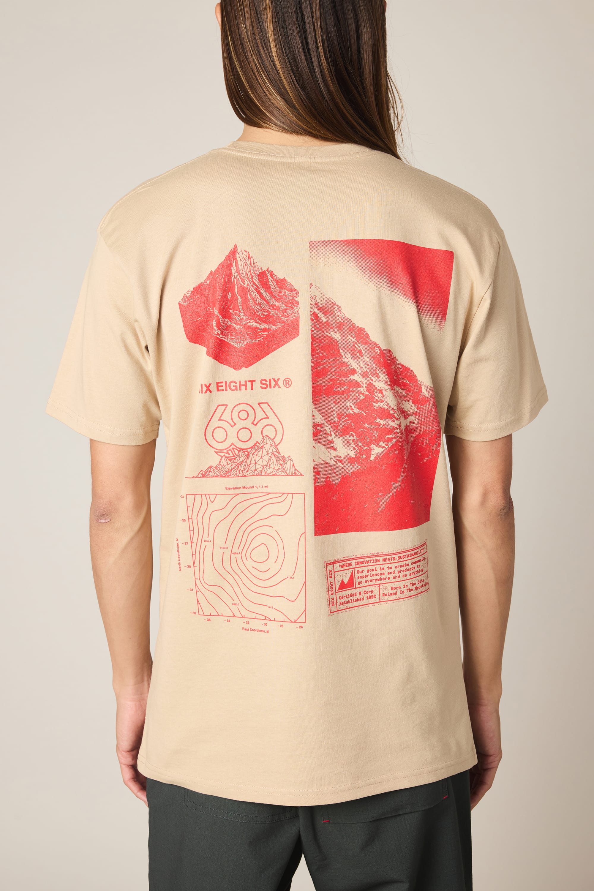 A person wearing the 686 Cross-Cut Authentic S/S Tee by 686— a beige men’s t-shirt featuring red mountain graphics, a contour map, and text on the back. Made from 100% cotton, the tee’s soft fabric is visible where the wearer’s long hair partially covers the collar. The background is plain and light.