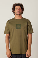 A man with curly brown hair wears the 686 Knockout Authentic S/S Tee, an olive green 100% cotton men's t-shirt with a subtle 686 logo on the chest, standing facing the camera against a plain light background.