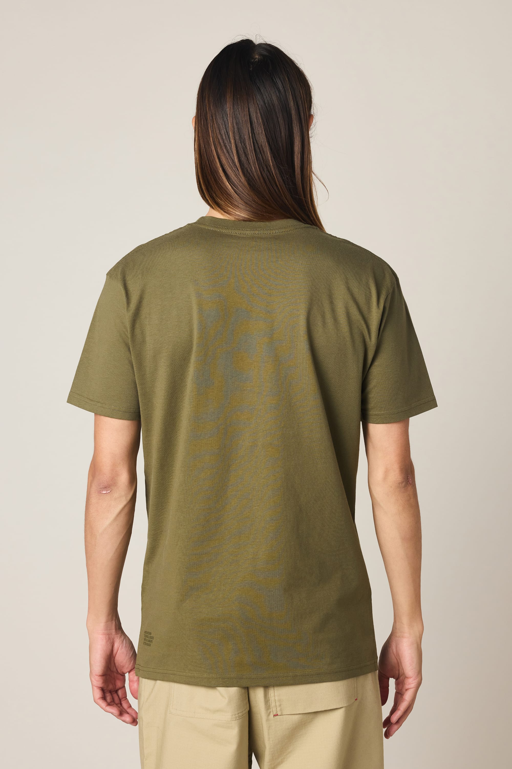 A person with long hair, viewed from behind, wears the 686 Knockout Authentic S/S Tee by 686 in olive green and beige pants, standing against a plain light background.
