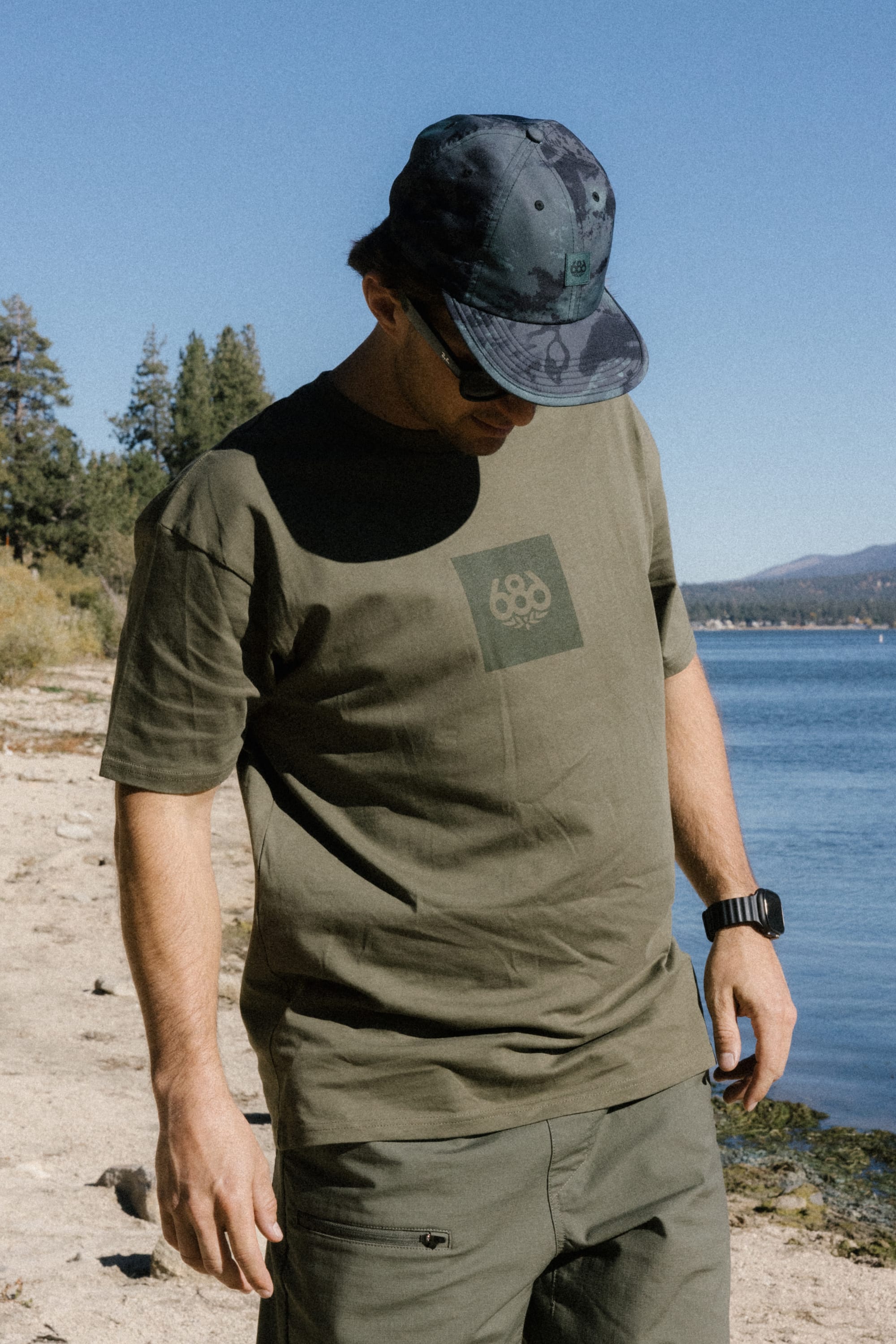 Wearing the 686 Knockout Authentic S/S Tee by 686 and a camouflage cap, a person stands on a lakeshore, looking down, with trees and mountains in the background under a clear blue sky.
