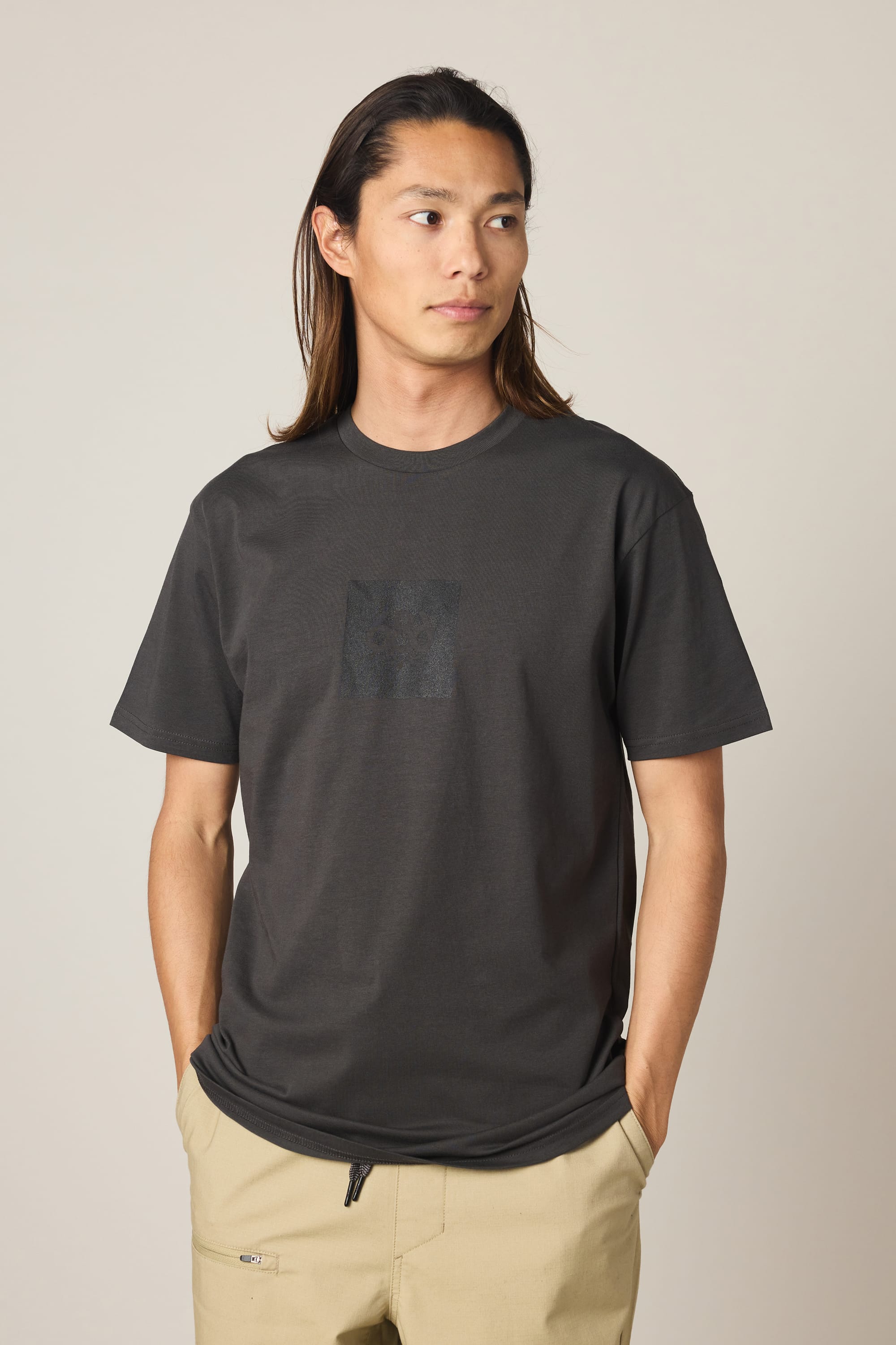 A person with long hair wears the 686 Knockout Authentic S/S Tee in dark gray, paired with beige pants, standing against a plain light background with hands in pockets, looking to their left.