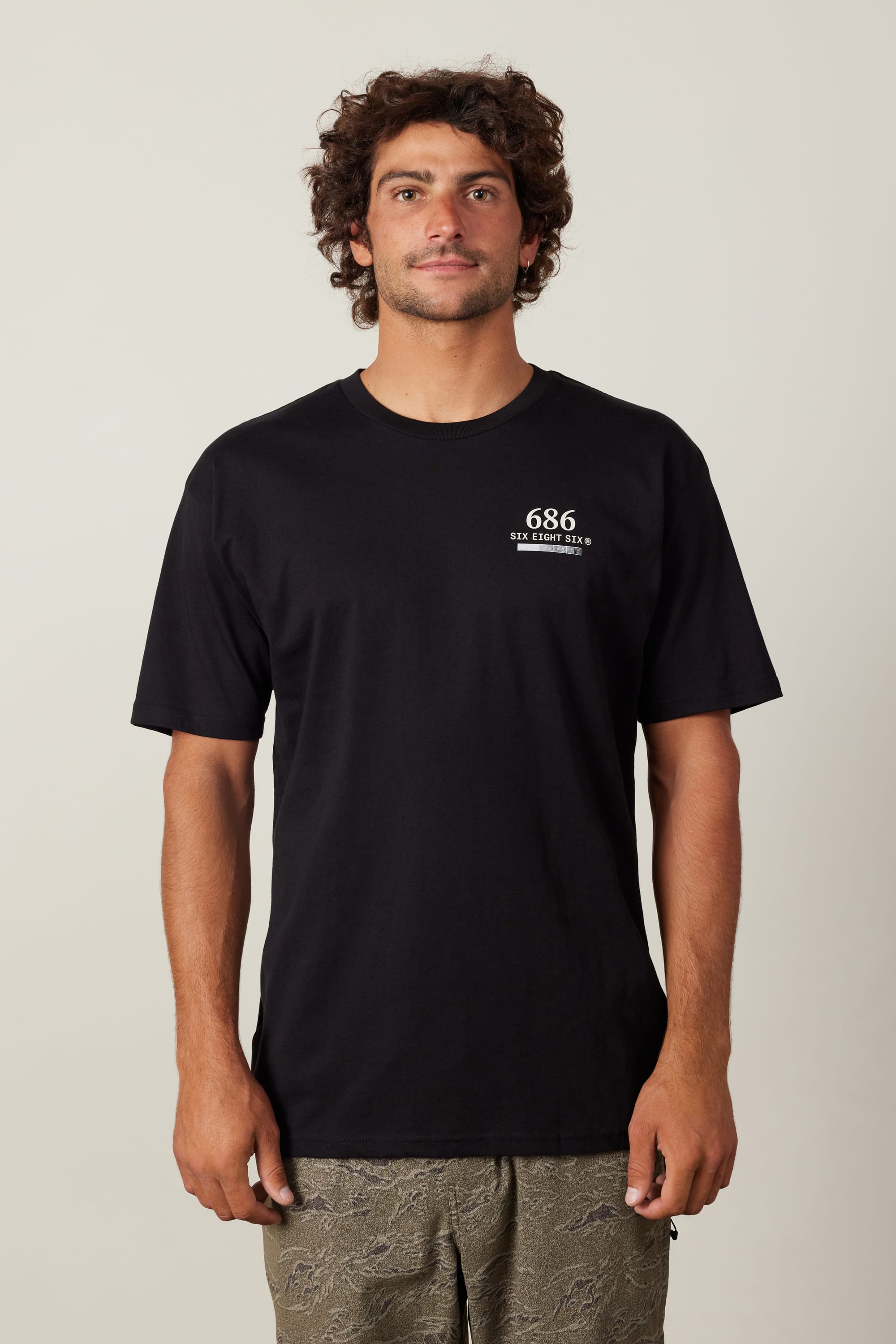 A man with curly hair wears the 686 Path Authentic S/S Tee, a black 100% cotton men’s t-shirt with “686” and white text on the left chest, paired with patterned khaki shorts. He stands facing forward against a plain light background.