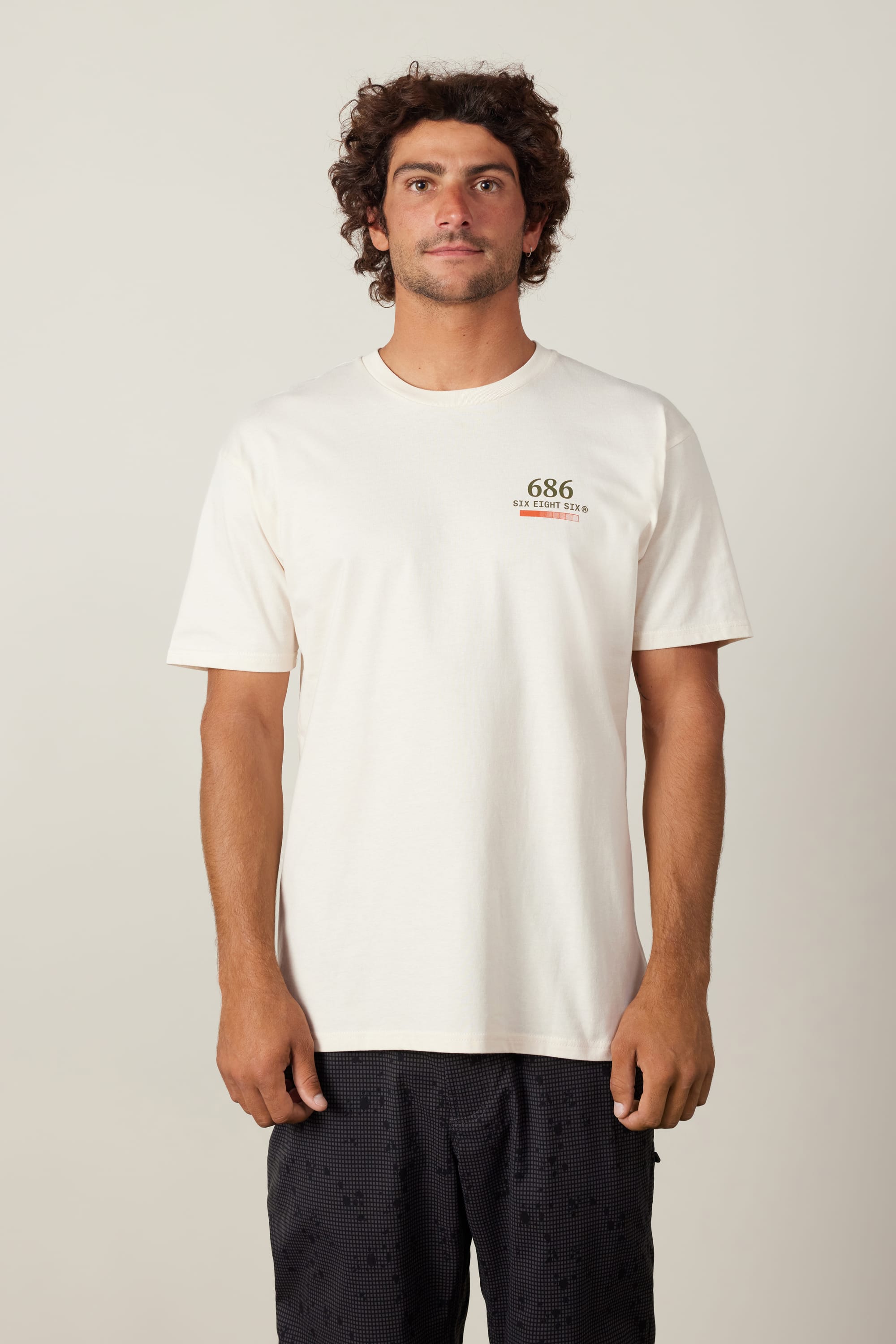 A man with curly hair wears the 686 Path Authentic S/S Tee by 686, featuring "UNDEFEATED" text on the chest, paired with black shorts, standing against a plain light background.