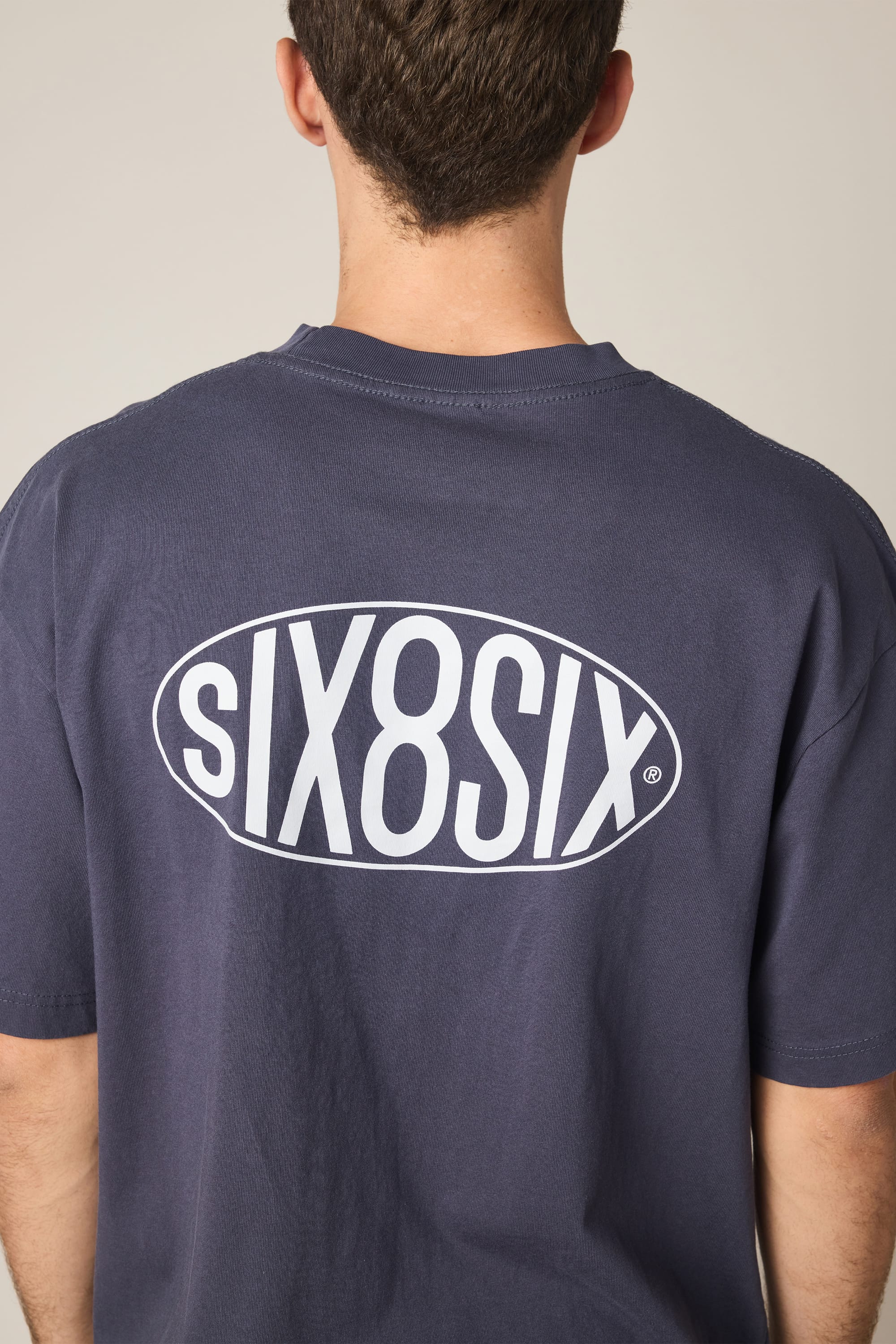 A person is shown from behind wearing the 686 92 Relaxed S/S Tee, a navy blue men's t-shirt from 686. The back of the 100% cotton shirt features a large white "SIX8SIX" logo printed inside an oval.