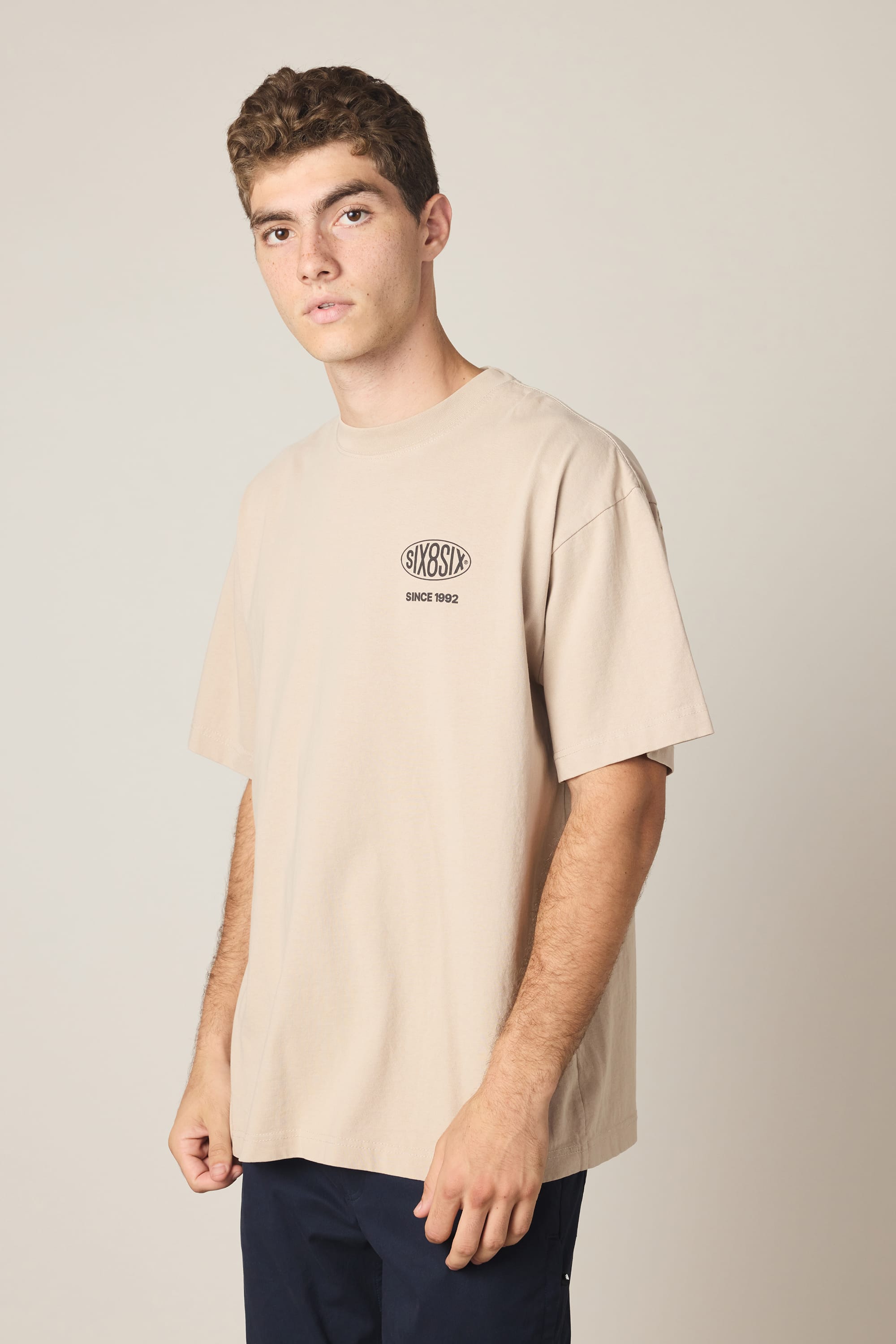 A young man with short curly hair wears the 686 92 Relaxed S/S Tee by 686—a loose, 100% cotton men’s T-shirt featuring a small graphic and text on the chest—paired with dark pants, standing against a neutral background.