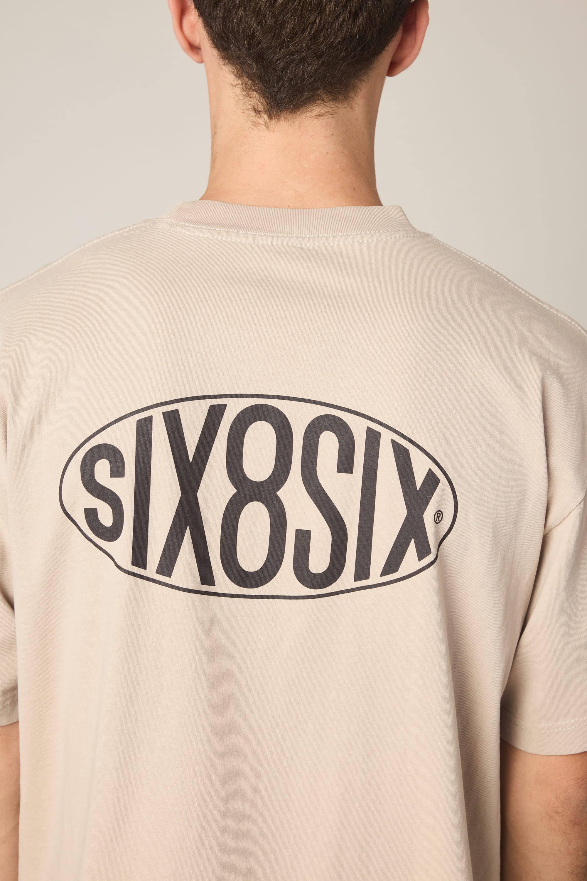 A person is seen from behind wearing the 686 92 Relaxed S/S Tee by 686, a beige men's t-shirt crafted from 100% cotton. The back of the shirt features bold black "sIX8SIX" lettering enclosed within an oval design, visible across the upper back and neck area.